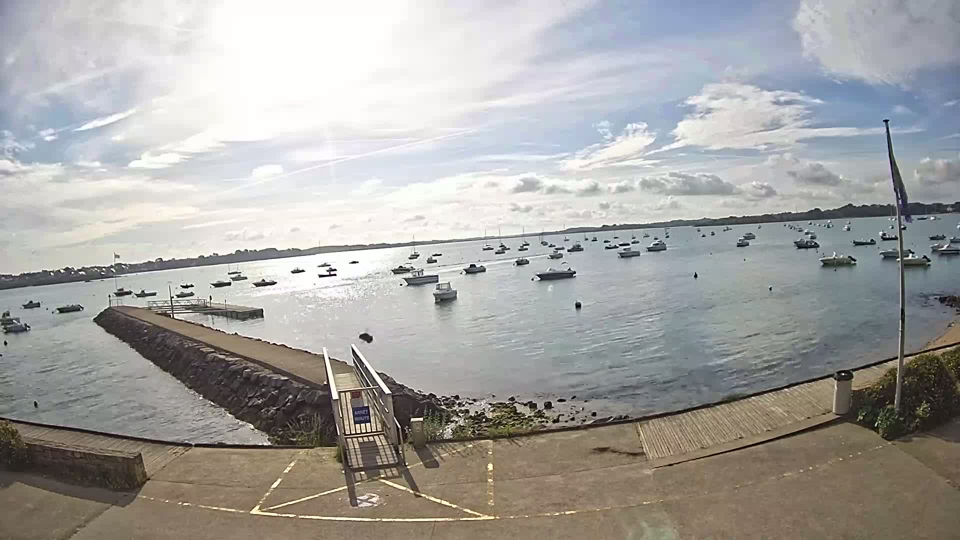 A sunny day with partly cloudy skies shows a calm body of water with numerous boats anchored, viewed from a stone jetty with a ramp leading to a paved area.