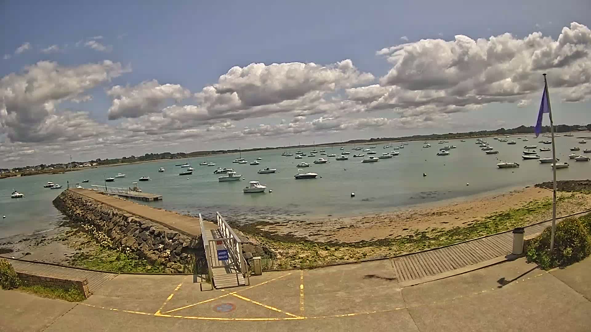 A calm, partly cloudy day reveals a harbor filled with numerous small boats, a stone jetty leading to a small beach, and a wooden walkway.