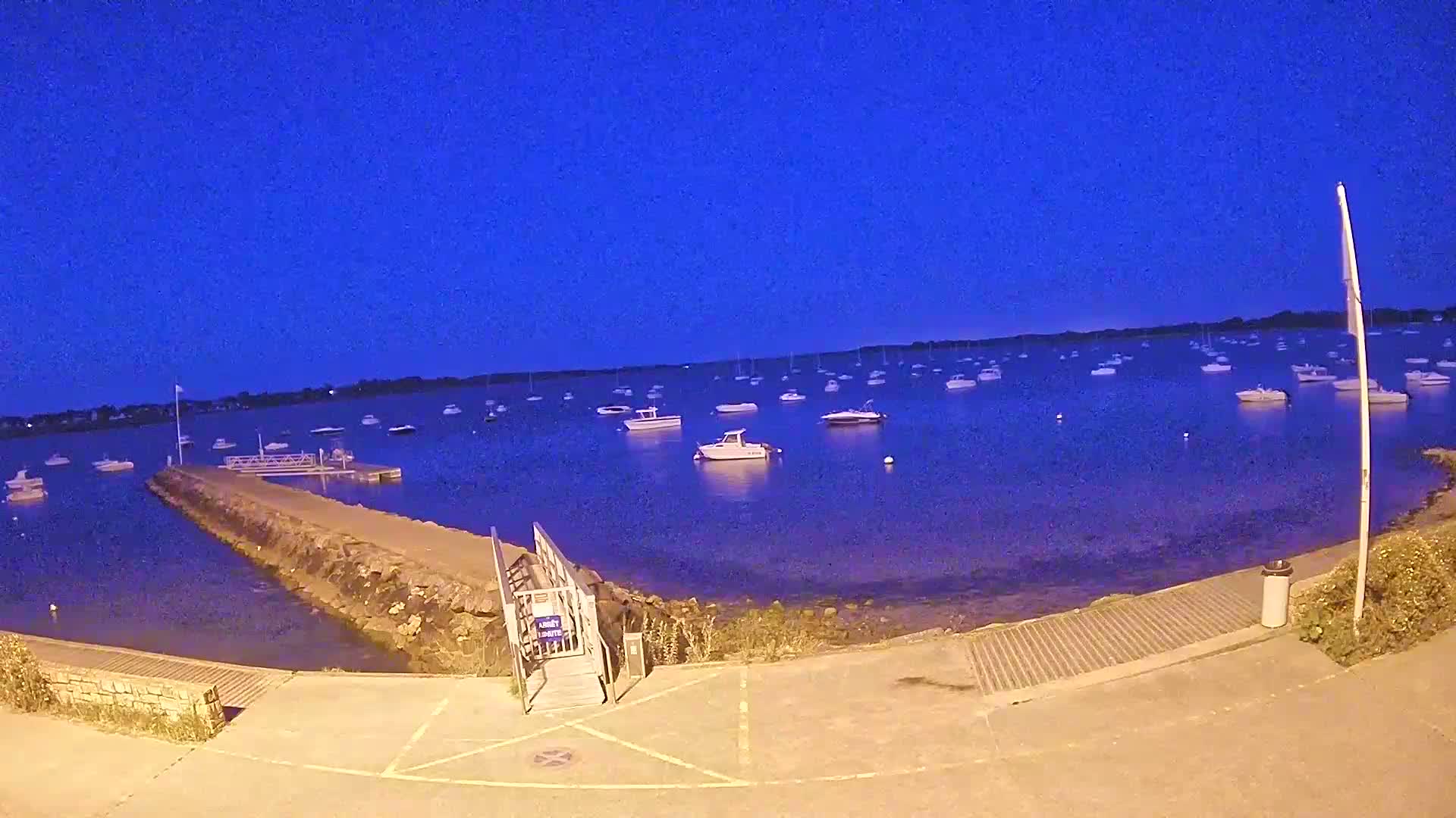 Under a dark blue twilight sky, numerous boats are moored in calm water near a small, paved waterfront area with a ramp leading down to the water.