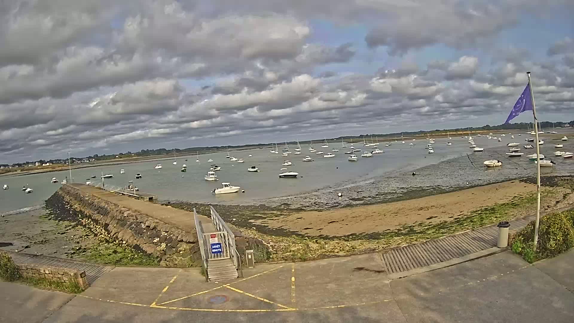 A mostly cloudy day reveals a calm bay with numerous boats at anchor near a stone jetty and sandy shore.