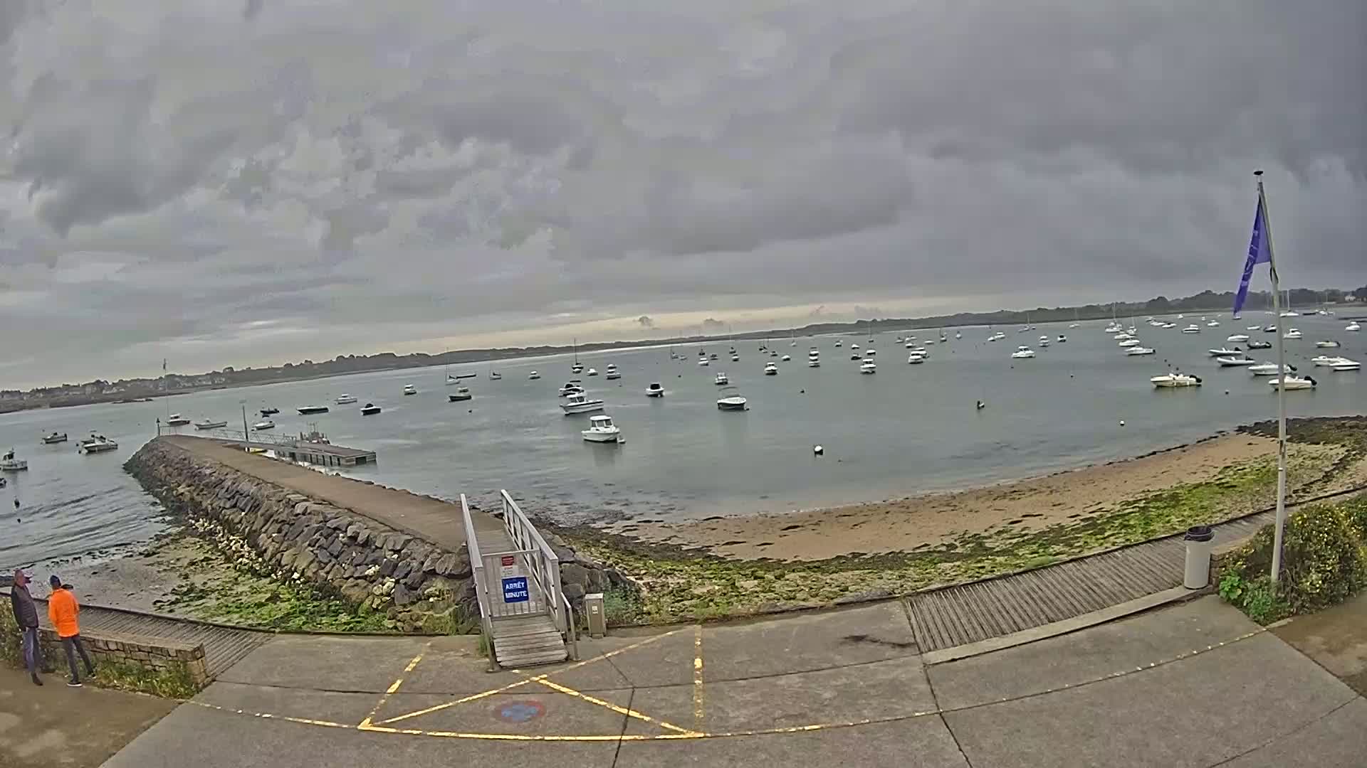A calm bay filled with numerous small boats under a cloudy sky, viewed from a paved area near a stone jetty.