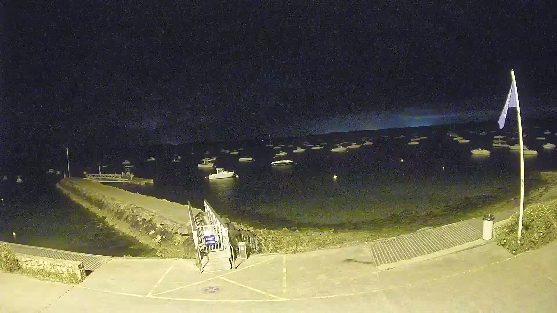 At night, numerous boats are moored in calm water near a stone seawall and a small boat ramp.