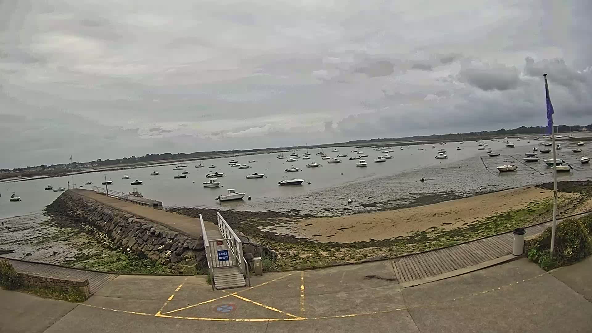 A calm bay under an overcast sky is filled with numerous small boats at low tide, with a stone jetty leading to a wooden ramp.