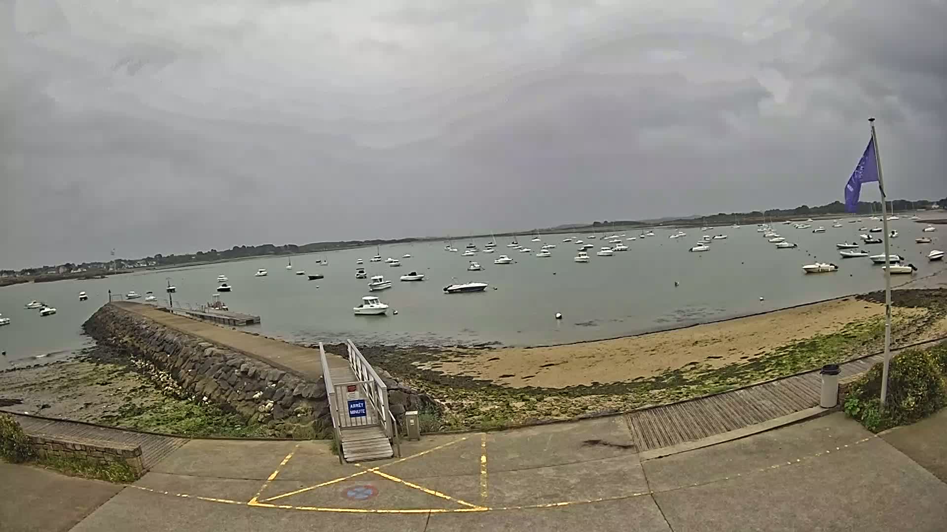A calm bay with numerous boats anchored under an overcast sky, viewed from a paved area leading to a stone jetty.