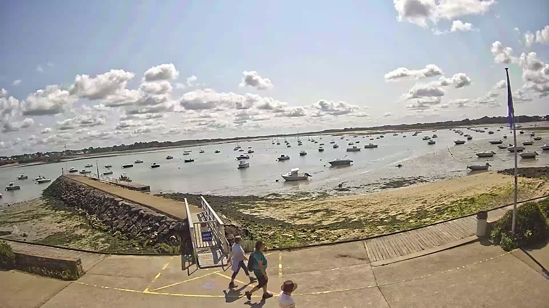 A sunny day with partly cloudy skies shows a calm body of water filled with numerous small boats, viewed from a paved area with a stone jetty, near which three adults and a child are walking.
