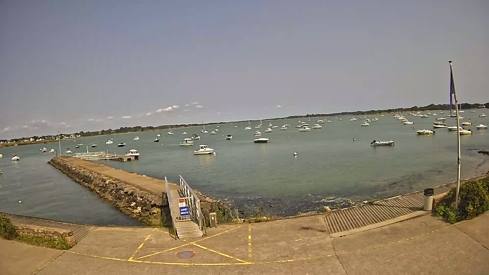A calm, sunny day shows a small harbor filled with numerous boats moored in shallow, green-tinged water, with a stone jetty and a wooden ramp leading to a paved area.