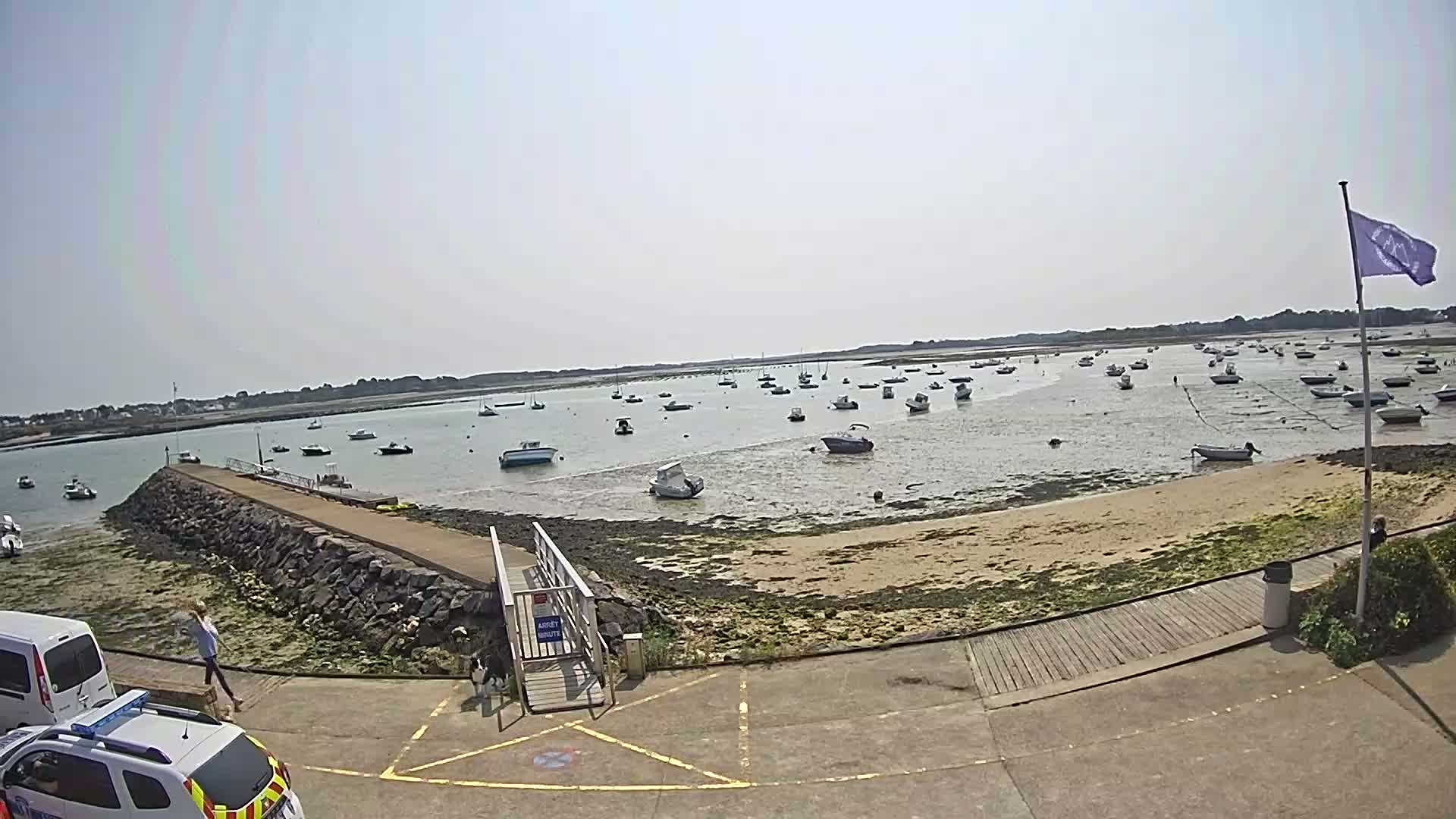A sunny day reveals a calm harbor filled with numerous boats at low tide, a stone jetty leads to a sandy shore.