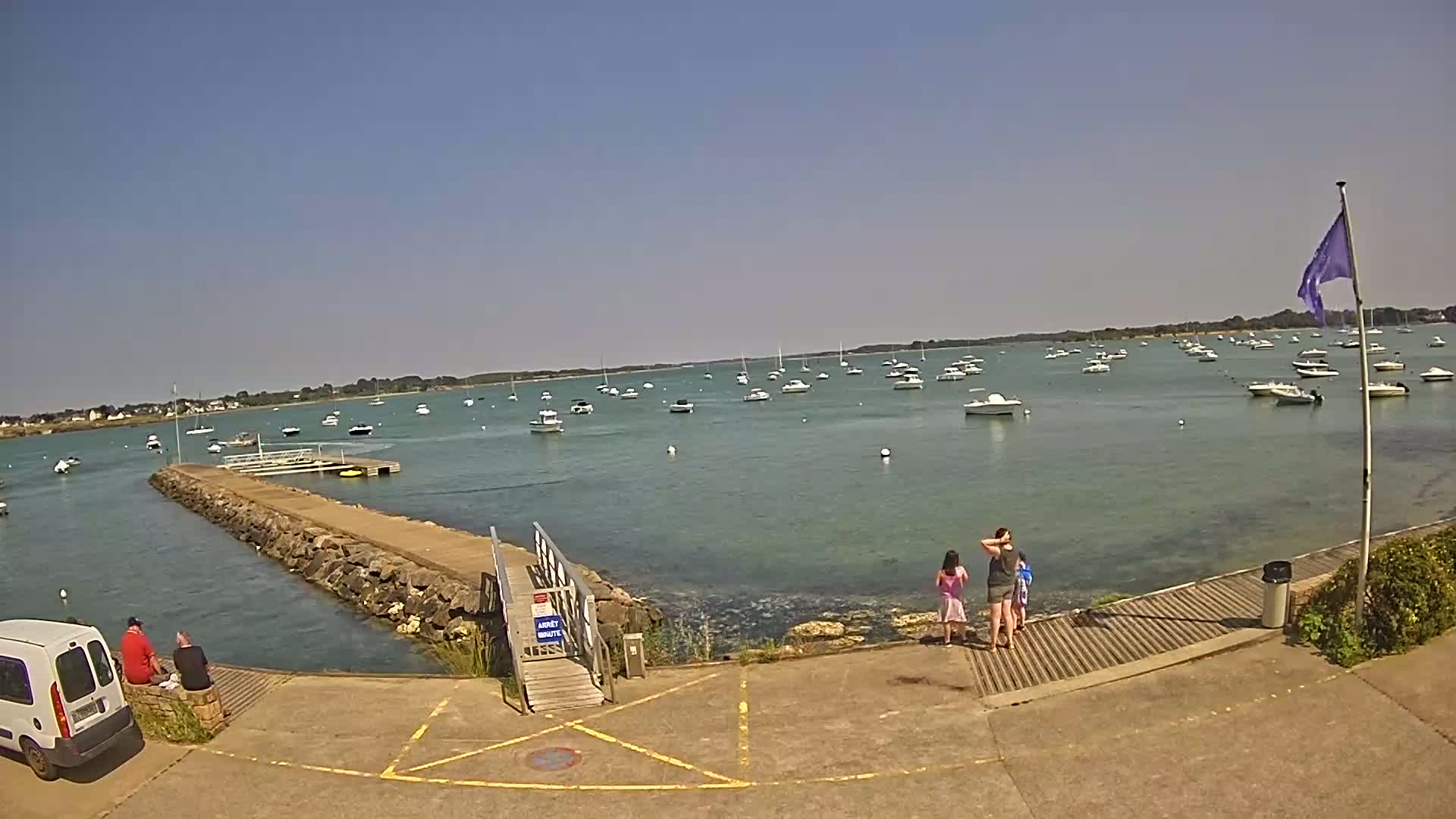 A sunny day at a harbor shows numerous boats at anchor, a stone jetty leading to a small ramp, and several people standing near the water's edge.