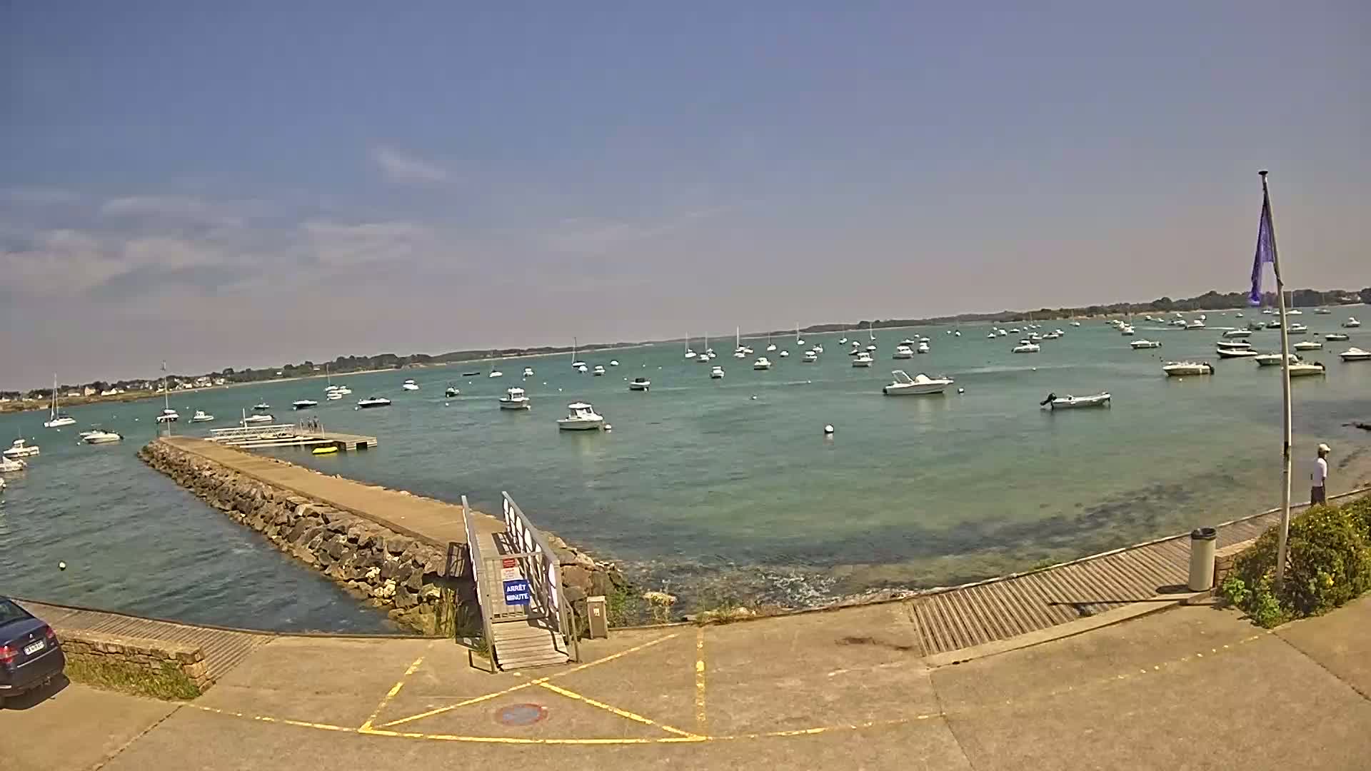 A sunny day at a harbor shows numerous boats at anchor in calm, clear water, with a stone pier leading to a small wooden gangway.