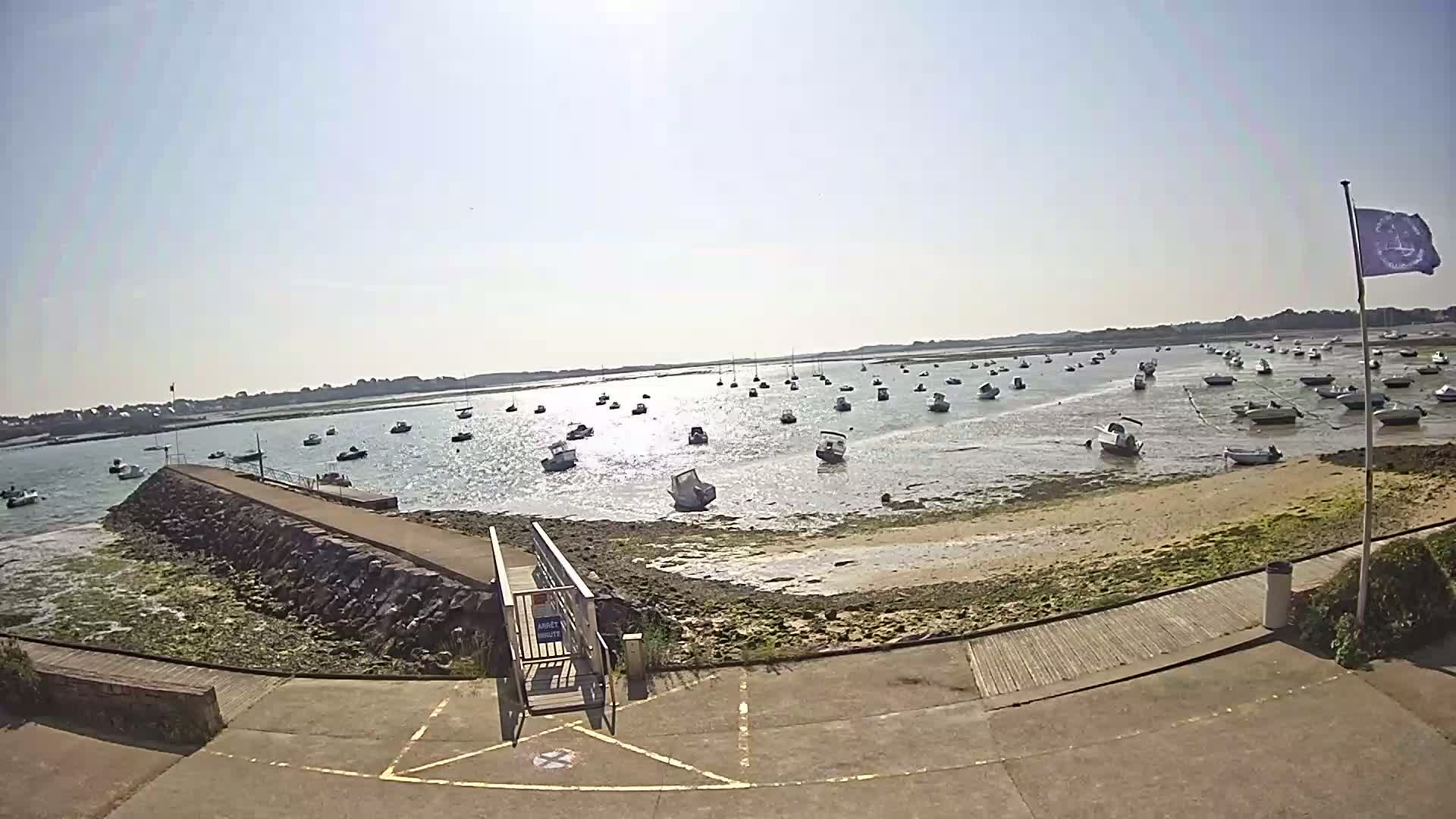 A sunny day reveals numerous small boats moored in a calm bay, with a stone jetty extending into the shallow, partially exposed shoreline.