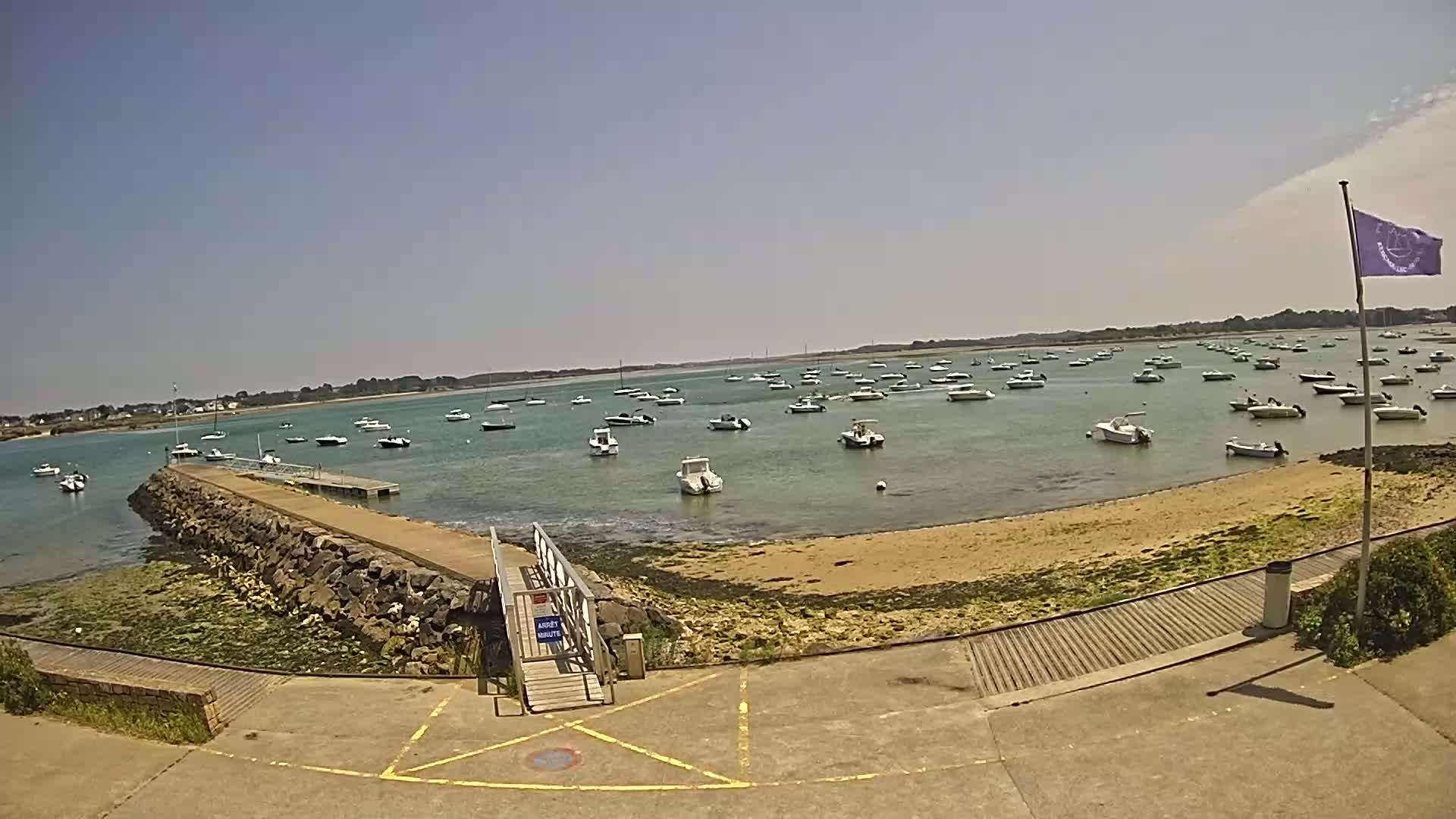 A sunny day shows a calm body of water with numerous small boats moored near a rocky shoreline and a small pier with a ramp leading to the water.