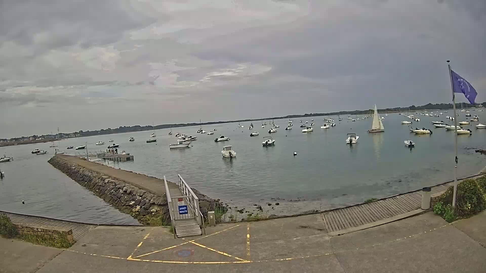 A calm, overcast day reveals numerous small boats and a sailboat anchored in a bay, viewed from a stone jetty with a wooden ramp leading to a paved area.