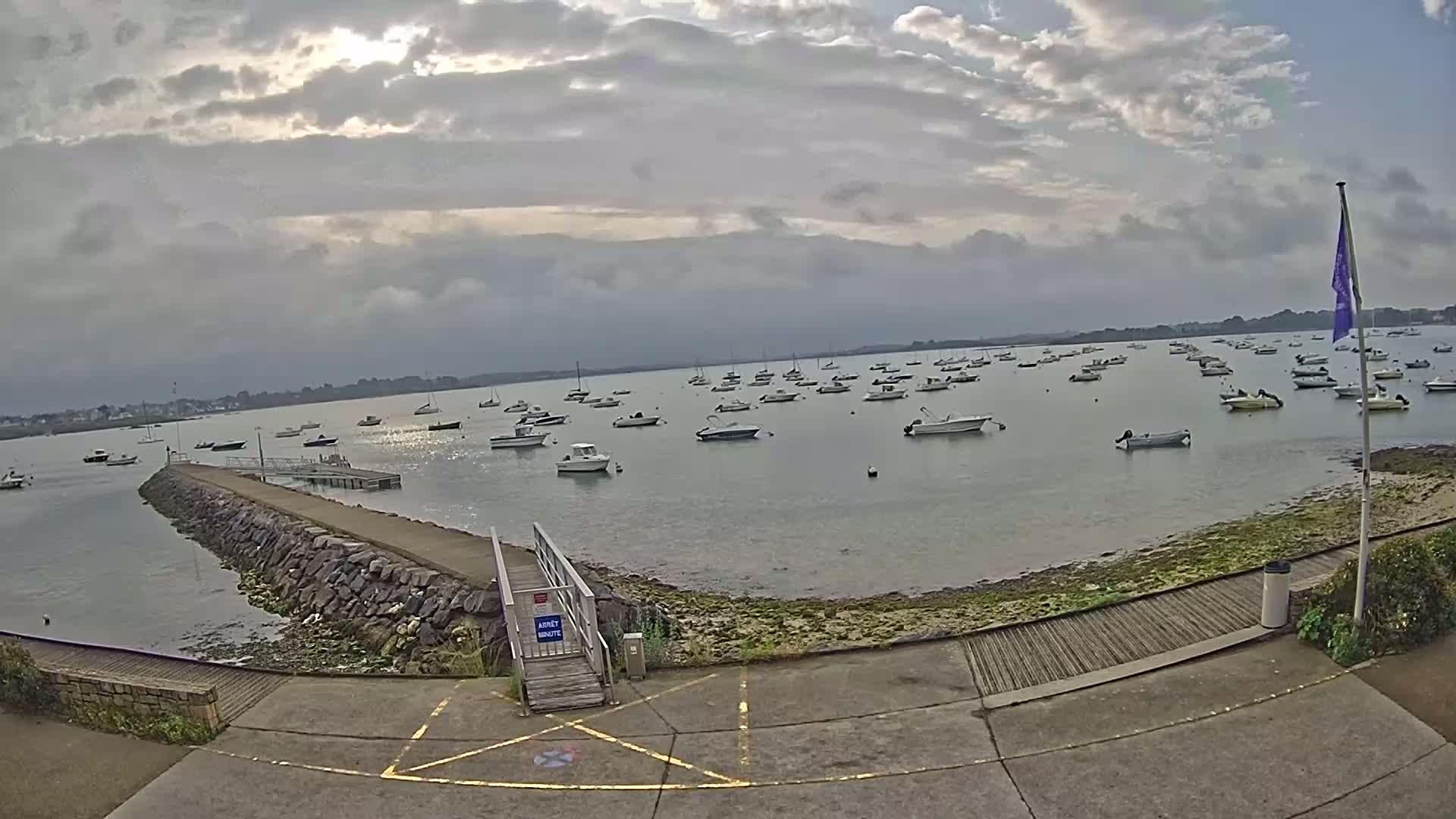 A calm bay with numerous small boats moored, viewed from a paved area with a stone jetty under a partly cloudy sky.