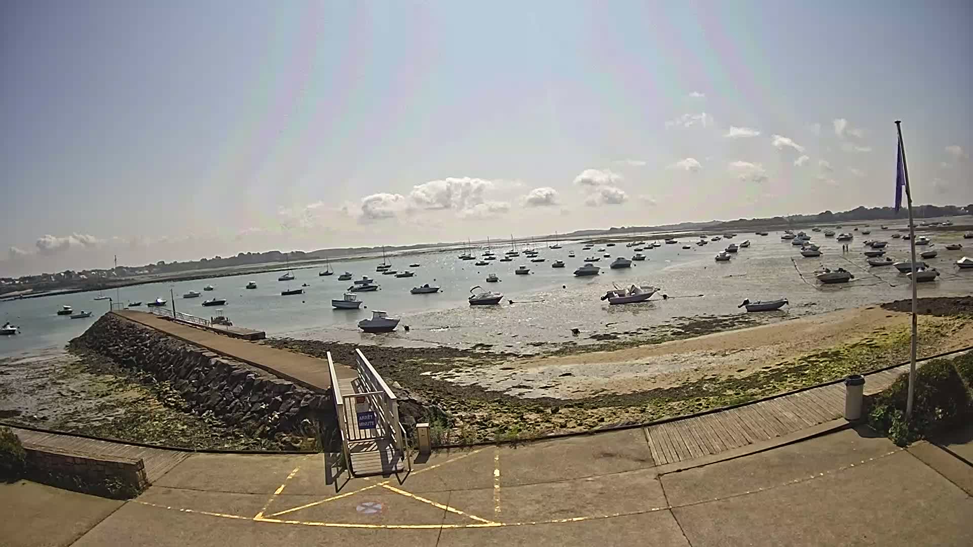 A sunny day reveals numerous boats at low tide in a shallow harbor, with a stone jetty and ramp leading to a paved area.