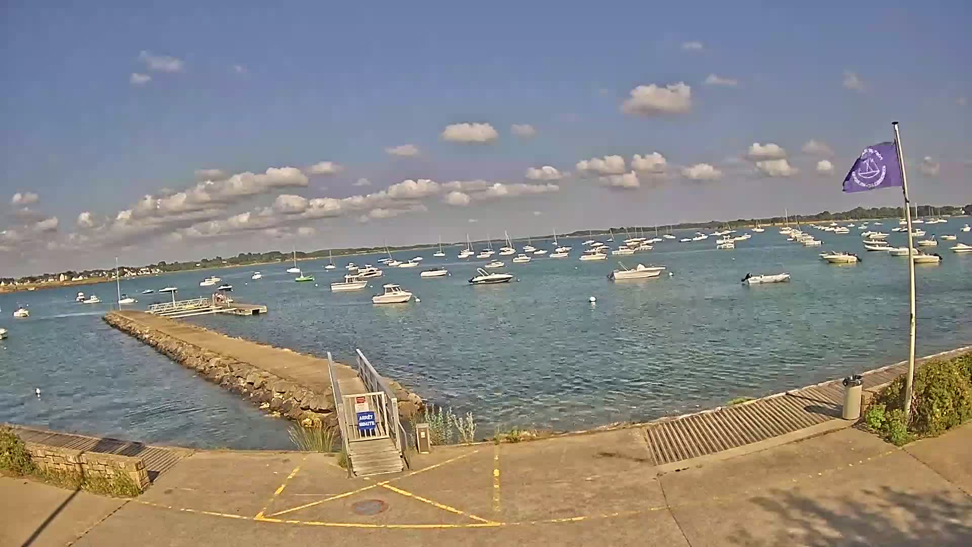 A calm, sunny day shows numerous boats moored in a bay, viewed from a paved waterfront area with a small pier and ramp.