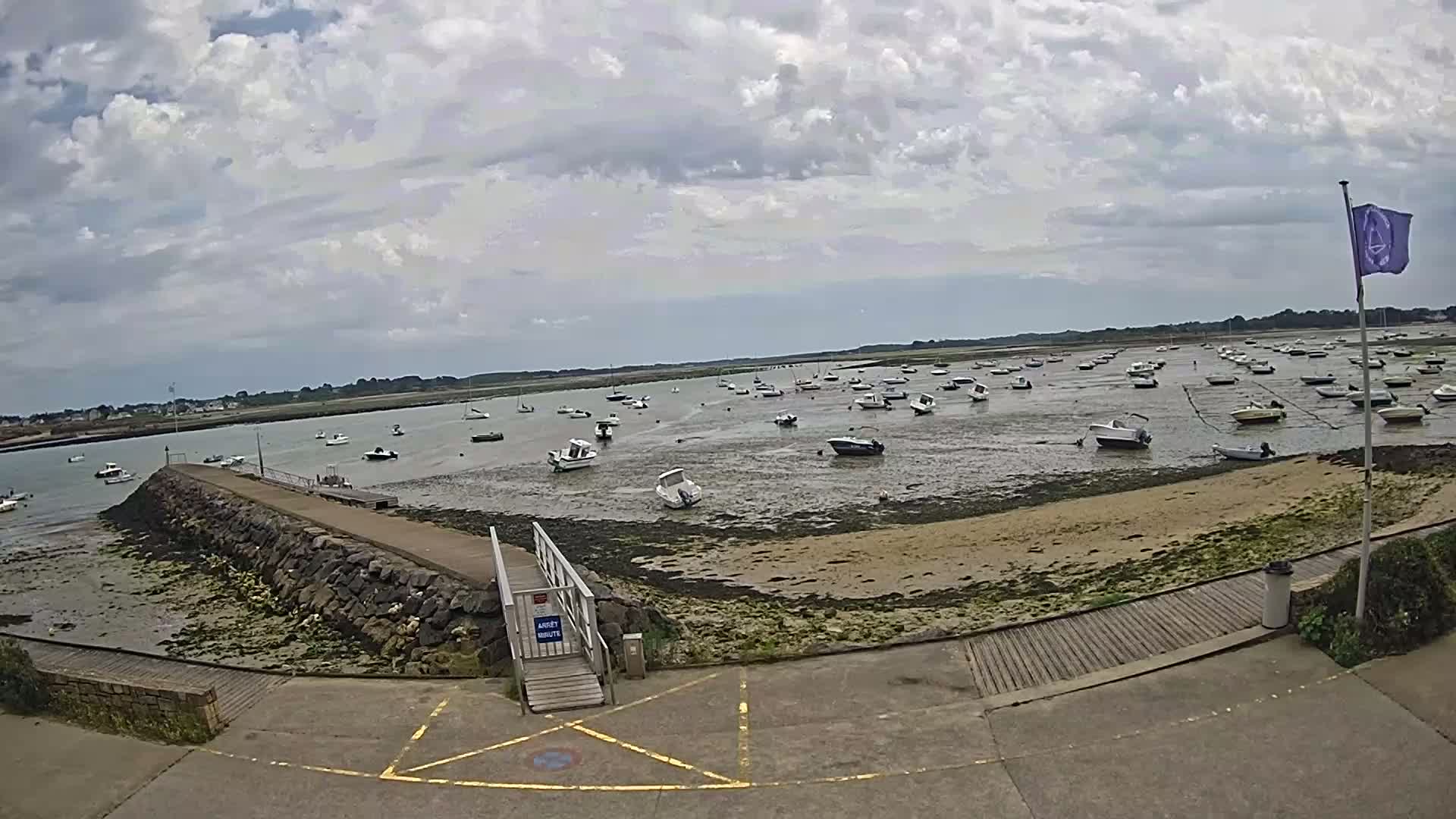 A mostly cloudy day reveals numerous small boats resting in shallow, sandy water near a stone pier and walkway.