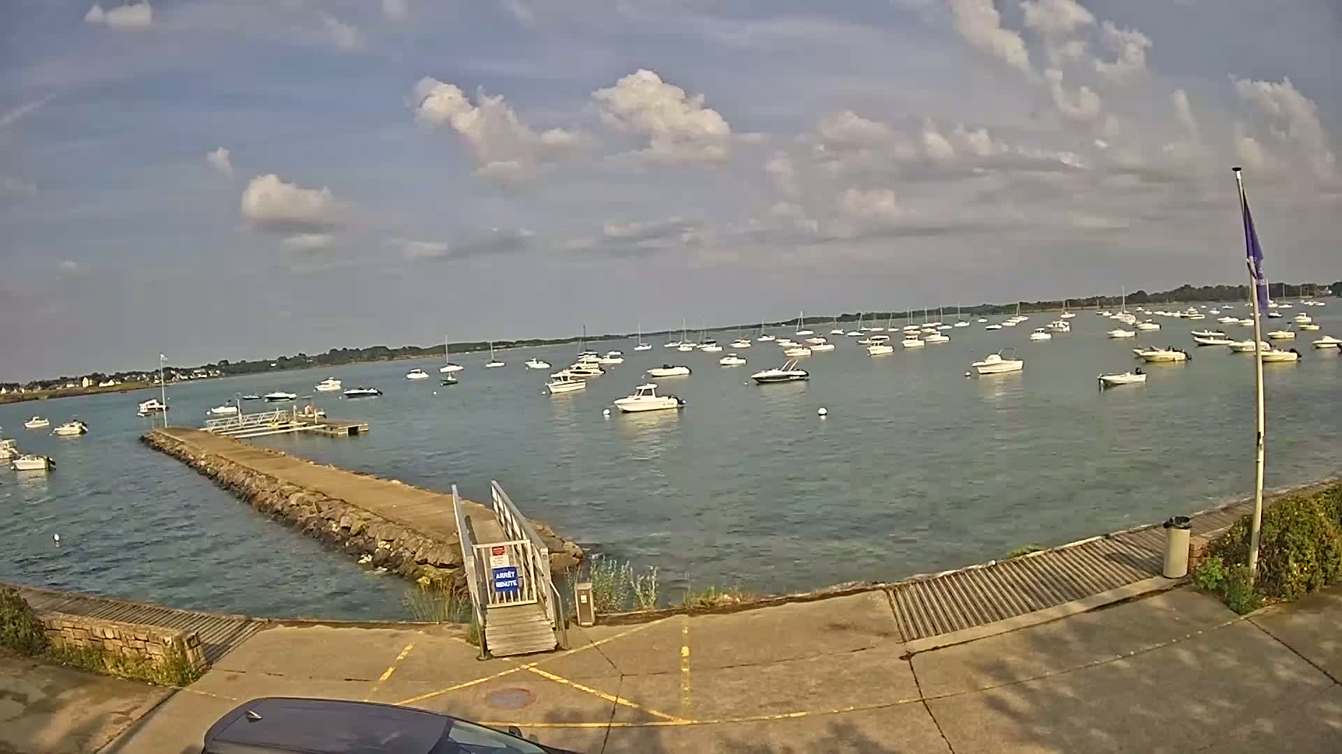 A calm, partly cloudy day shows numerous boats moored in a harbor, accessed by a stone jetty with a ramp.