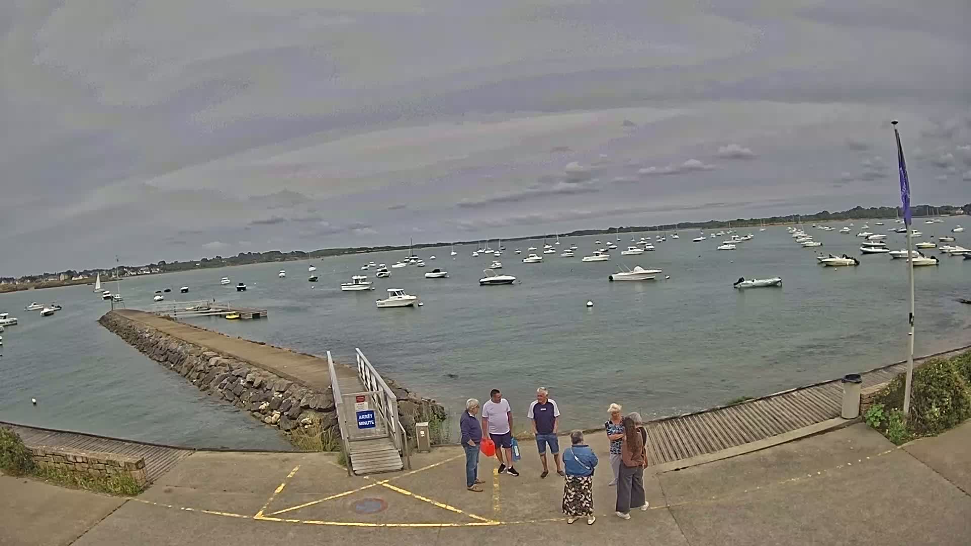 A group of people stands near a stone jetty overlooking a calm bay filled with numerous boats under a cloudy sky.