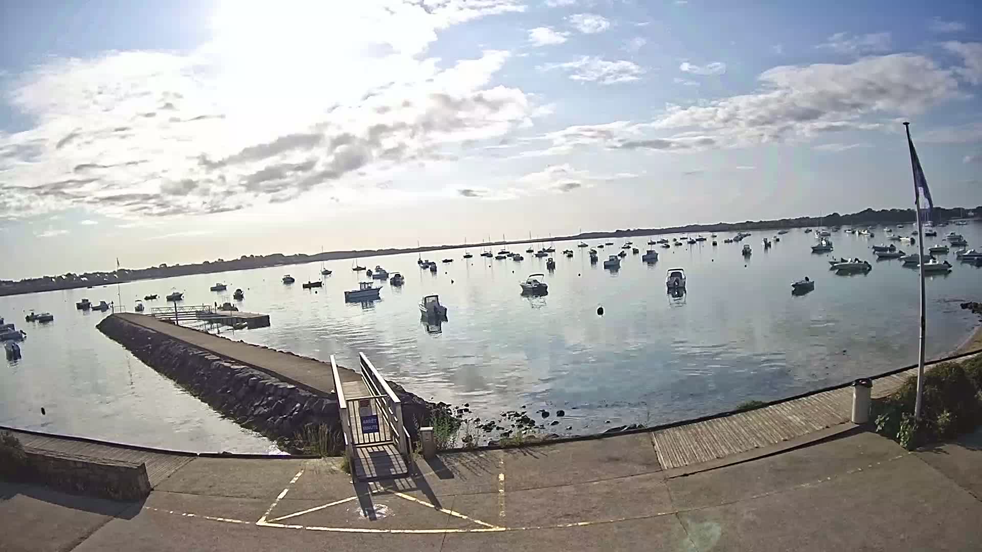 A calm, sunny day shows numerous small boats moored in a harbor, with a stone jetty leading to a small ramp.