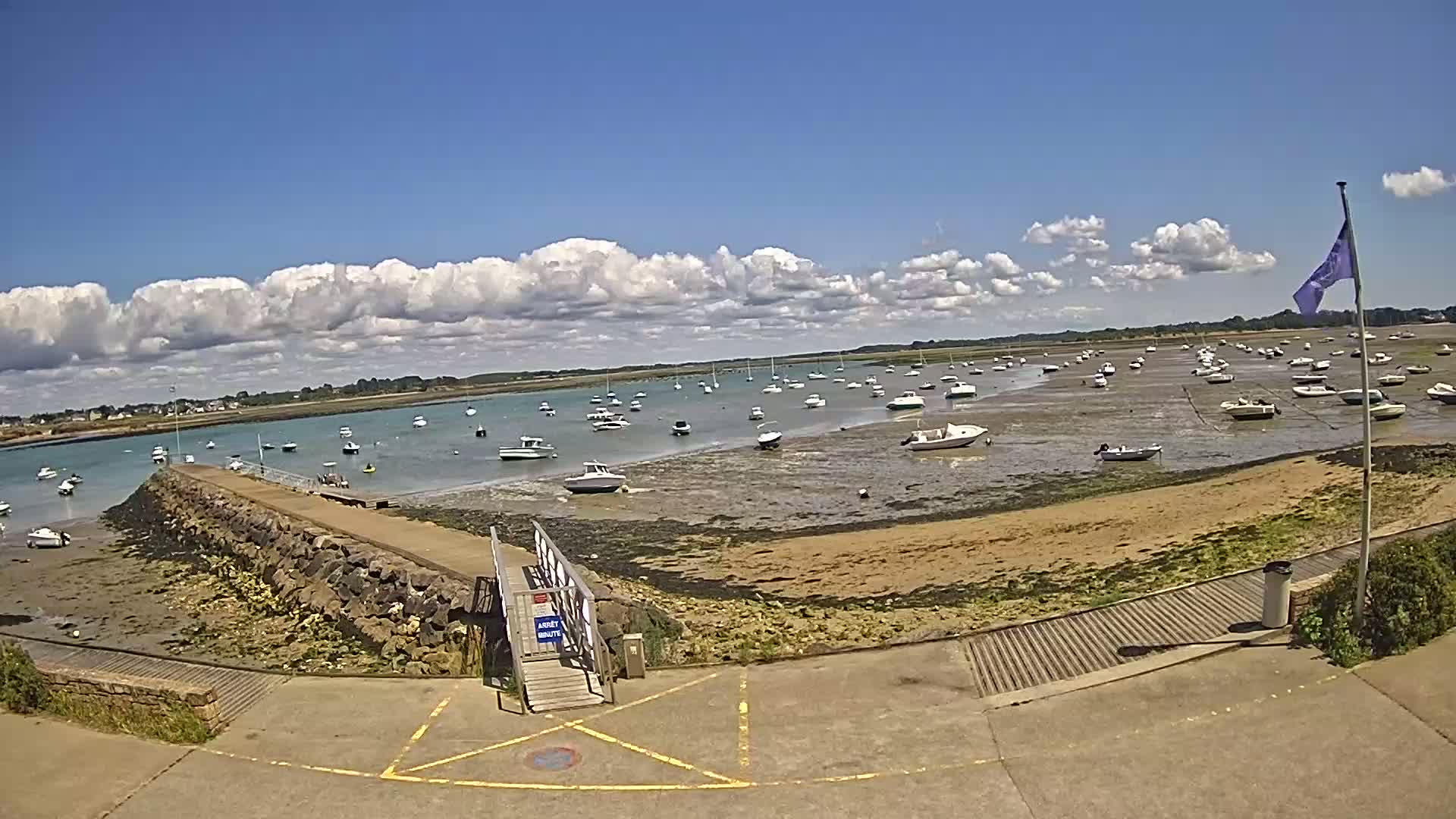 A sunny day with partly cloudy skies shows numerous boats resting on a tidal flat near a stone jetty and paved area.