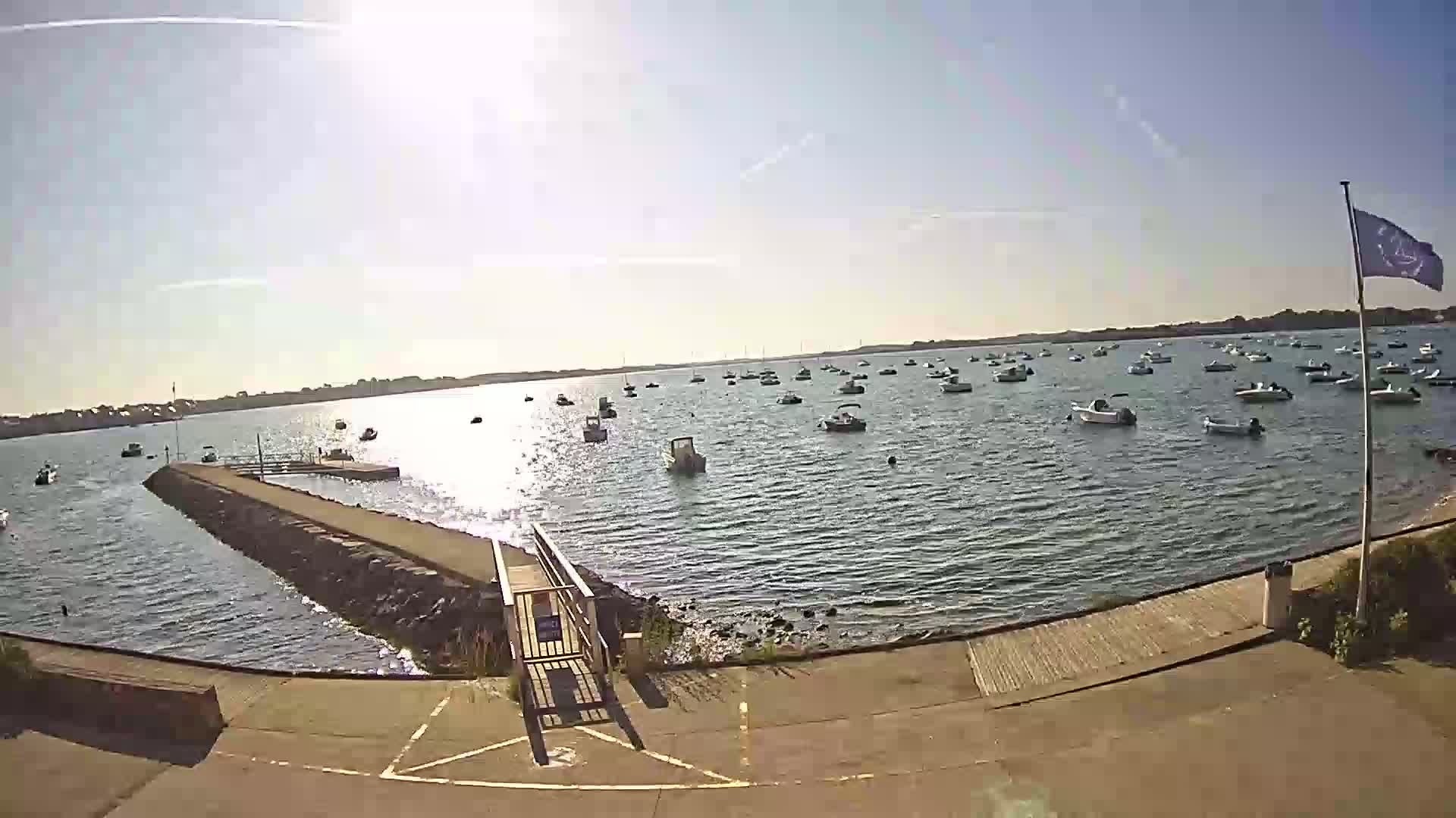 A sunny day shows numerous small boats moored in calm water, viewed from a paved boat ramp leading to a small stone jetty.