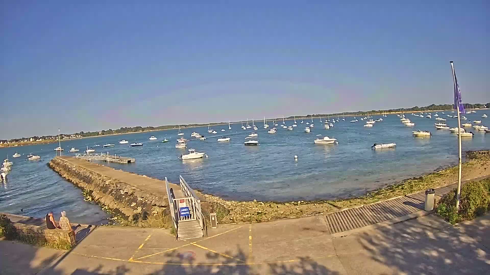 A calm, sunny day reveals a harbor filled with numerous boats, a short stone jetty leading to a small wooden ramp, and two people seated on a low wall.