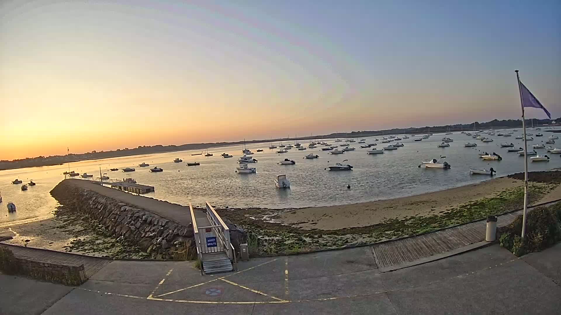 Many small boats are moored in calm water at sunset near a stone jetty and sandy shore.