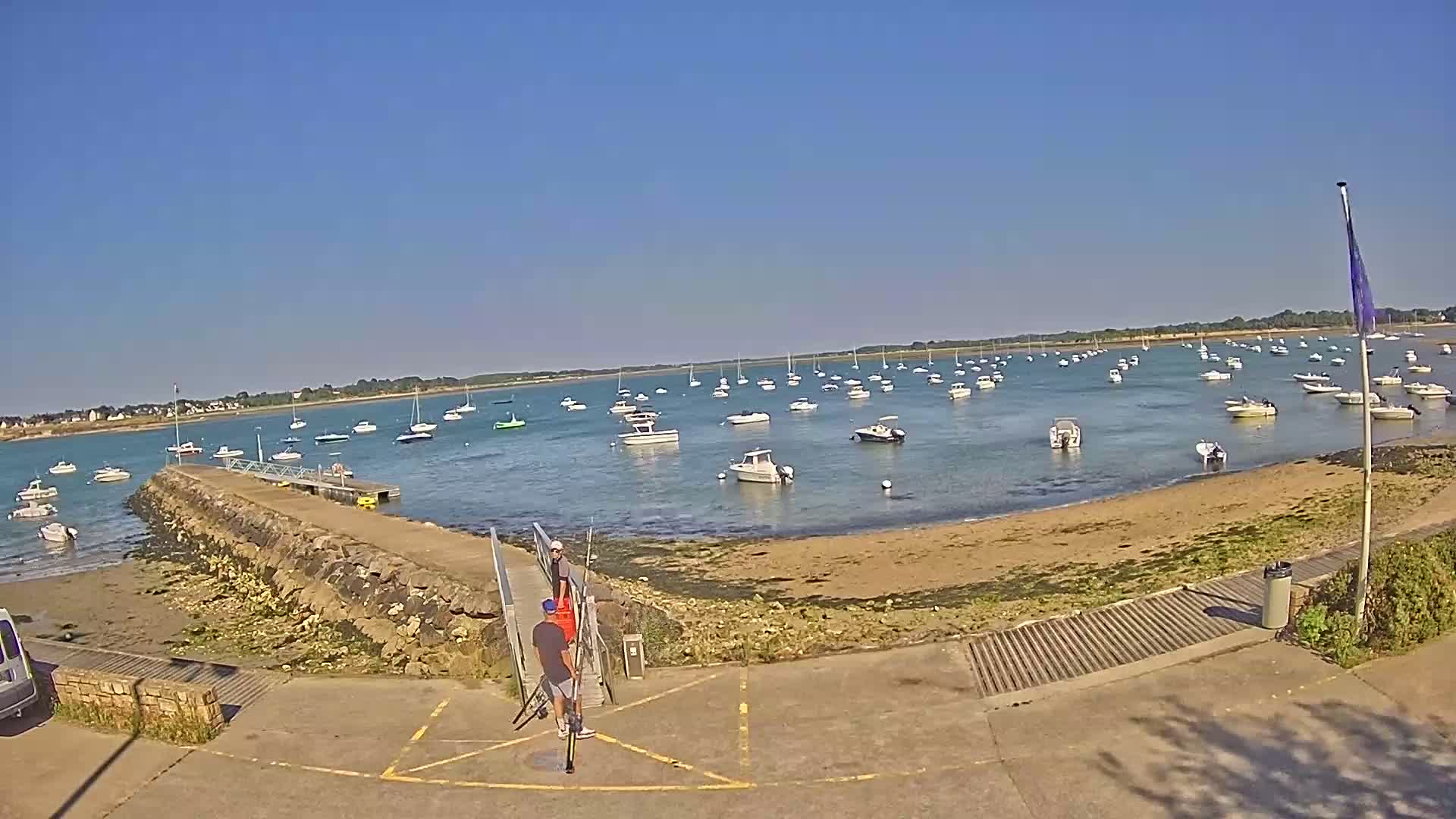 A sunny day at a harbor shows numerous small boats anchored in calm water, with a stone jetty and a few people near a boat ramp.