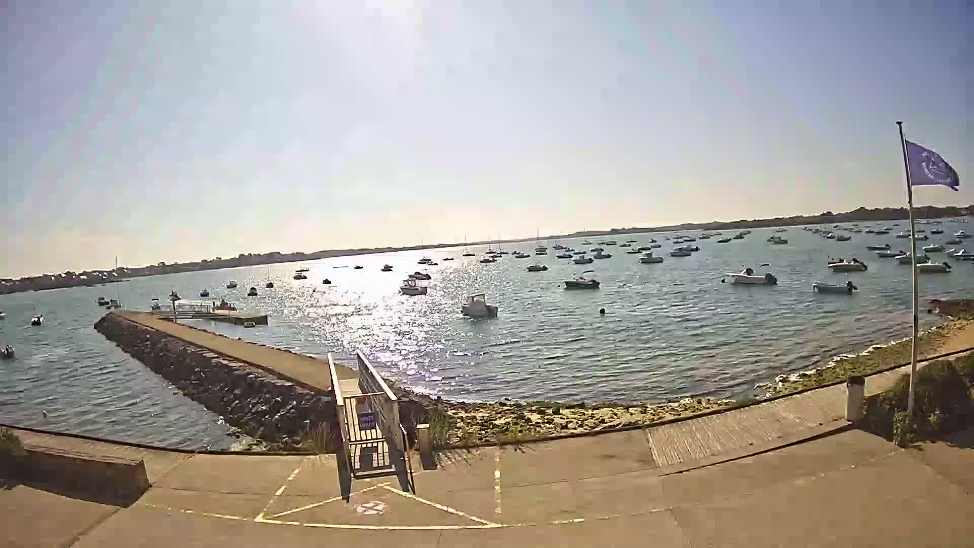 A sunny day shows numerous boats anchored in calm water, viewed from a paved area leading to a stone jetty.