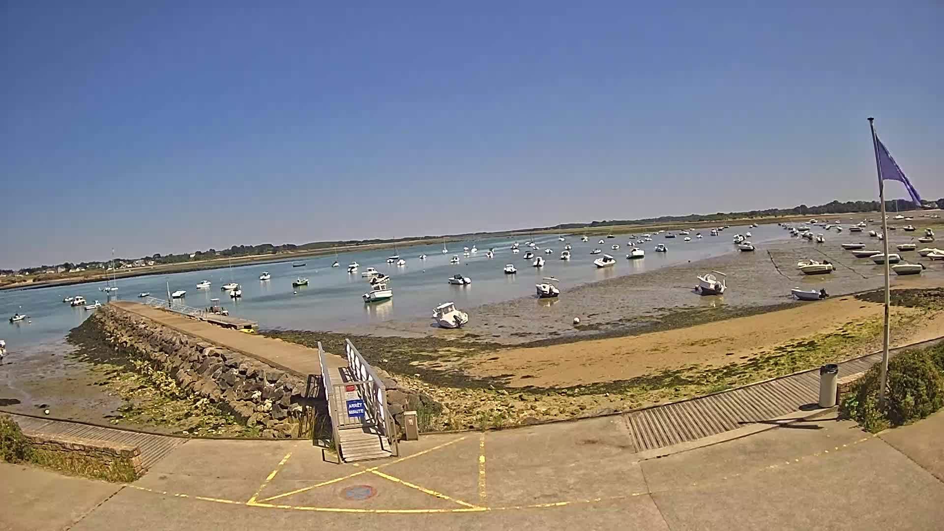 A sunny day reveals numerous small boats resting in shallow, calm water near a stone jetty and sandy shore.