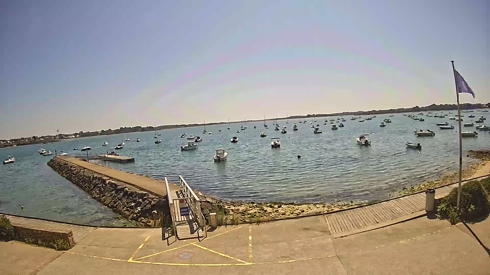 A calm, sunny day shows many small boats moored in a harbor, with a stone jetty leading to a wooden ramp.