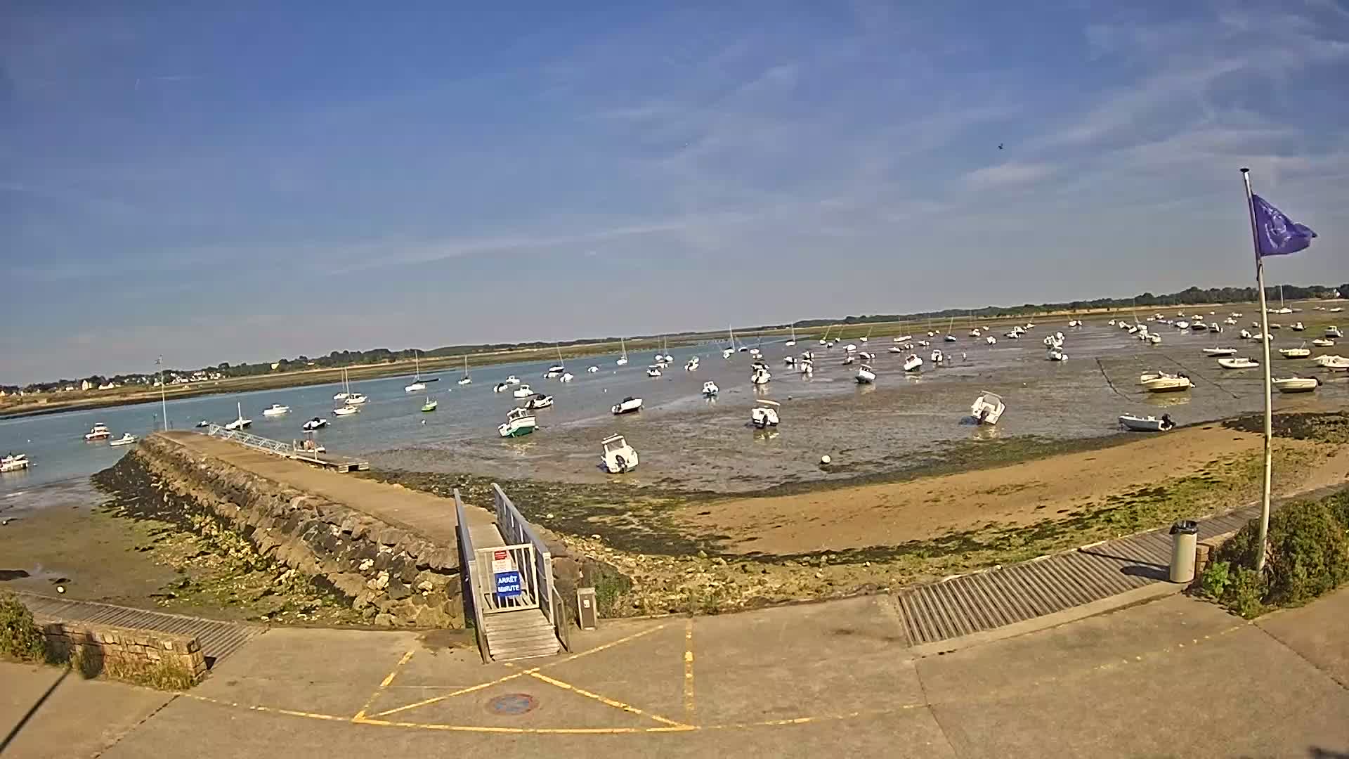 A sunny day reveals numerous small boats resting on a shallow, sandy seabed at low tide, near a stone jetty and ramp.