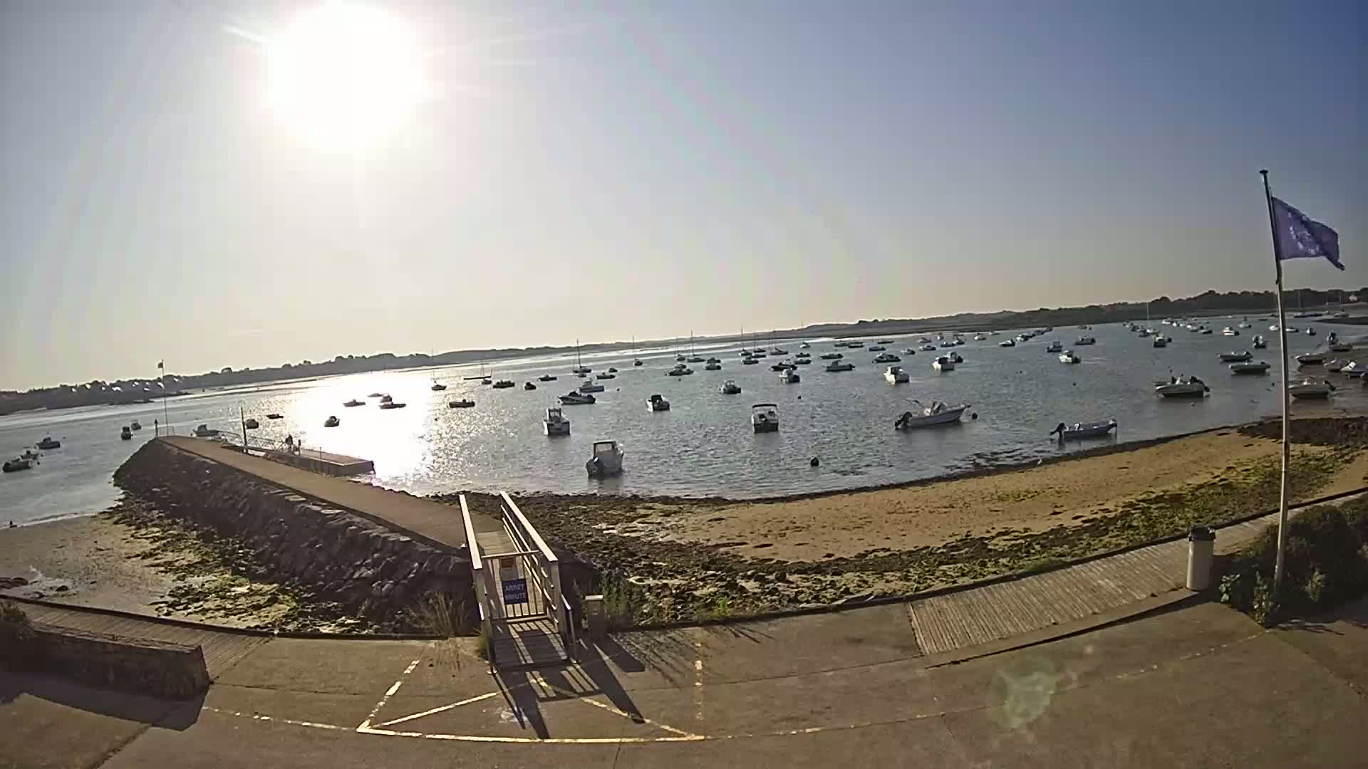 A sunny day reveals numerous boats moored in a calm bay, adjacent to a sandy beach and a stone jetty with a small access ramp.