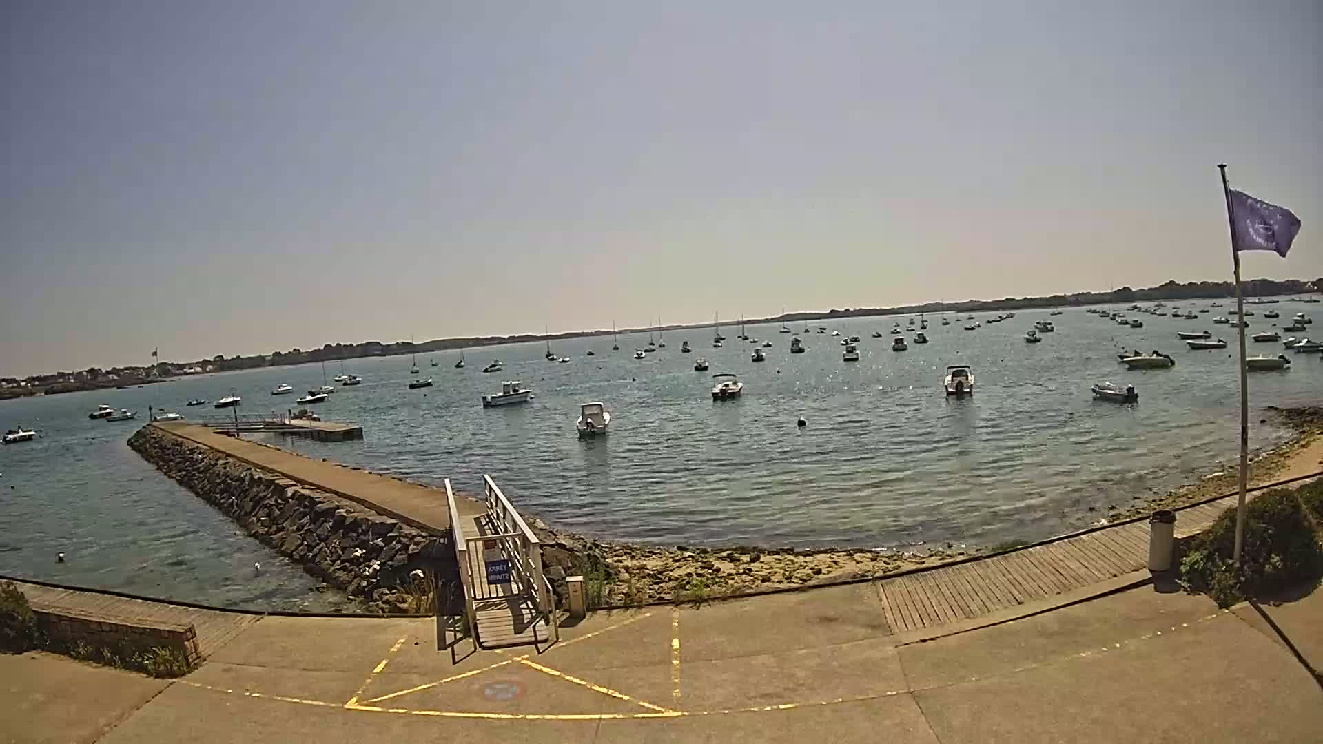 A calm, sunny day shows many small boats moored in a bay, with a stone jetty and wooden walkway leading to the water.