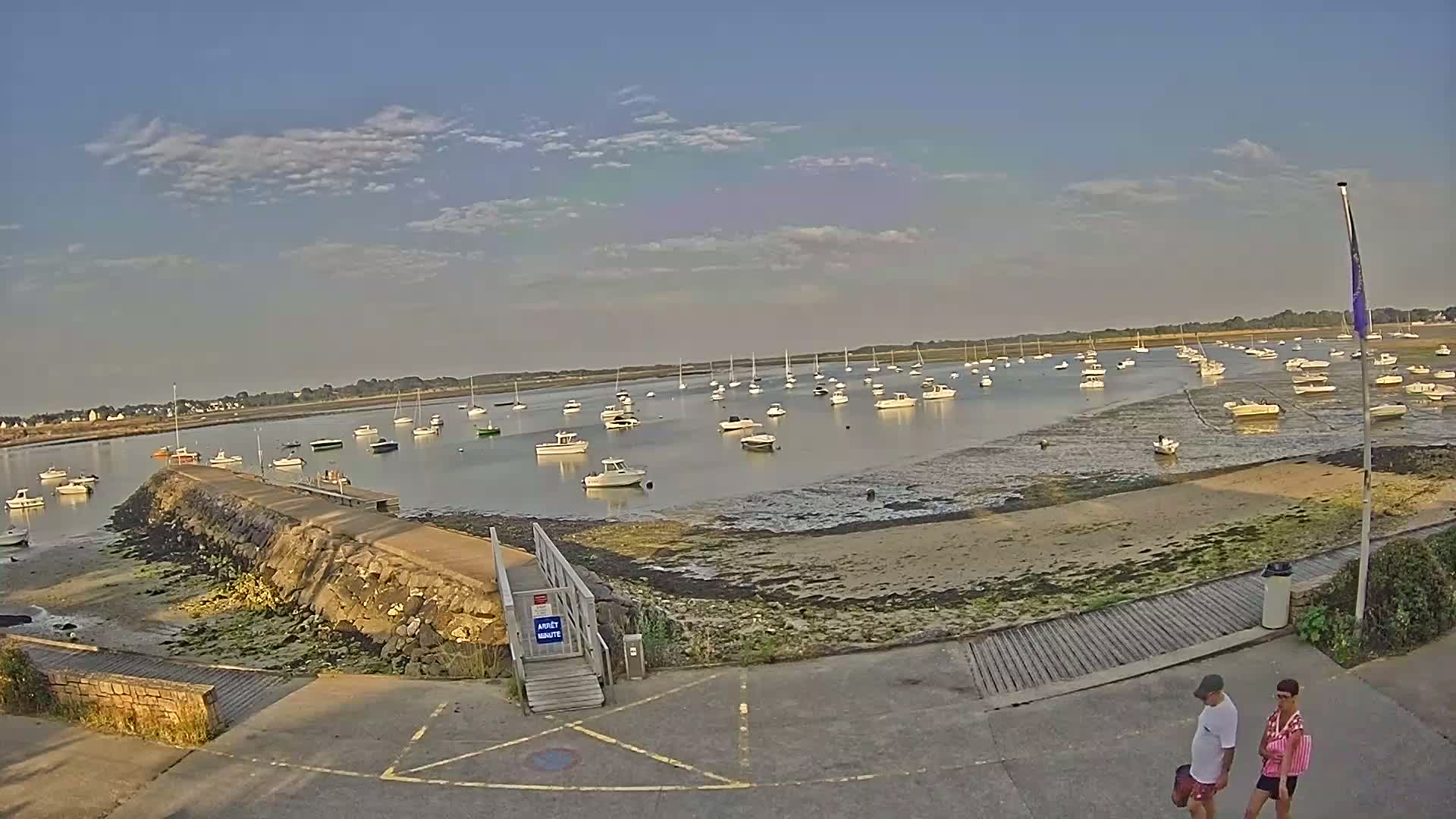 A calm body of water with numerous boats is visible at low tide under a partly cloudy sky, with two people walking near a stone jetty in the foreground.