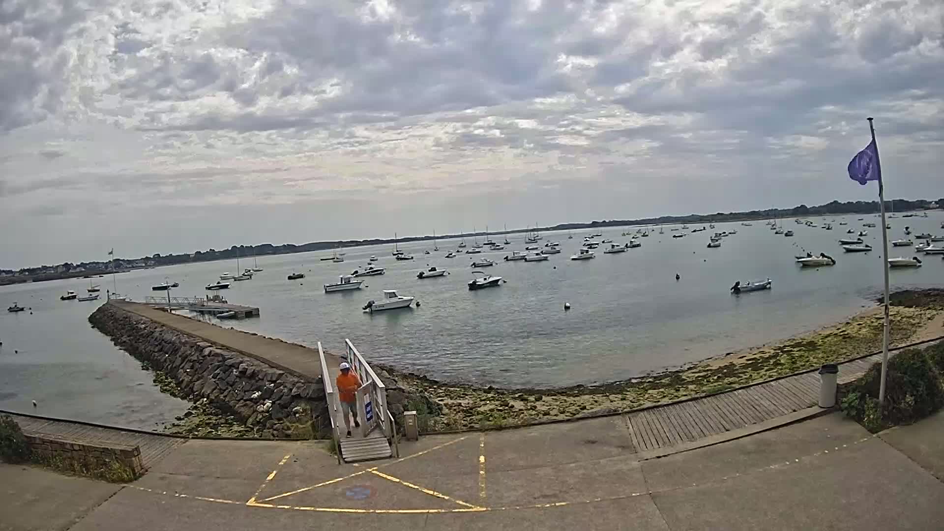 A calm body of water filled with numerous small boats is viewed from a stone pier on an overcast day.