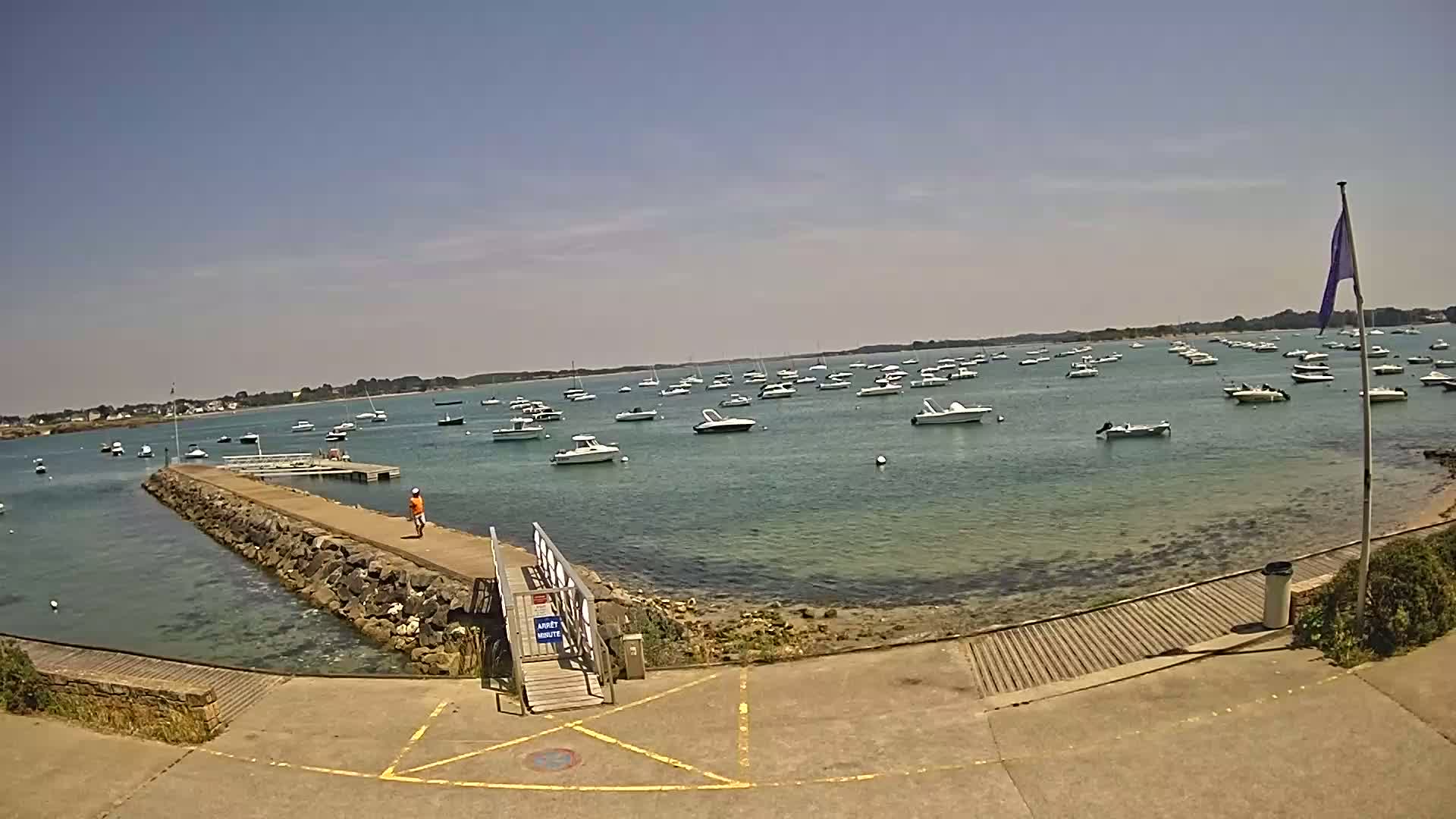 A sunny day at a harbor shows numerous boats at anchor in calm, clear water, with a stone jetty leading to a small wooden walkway.