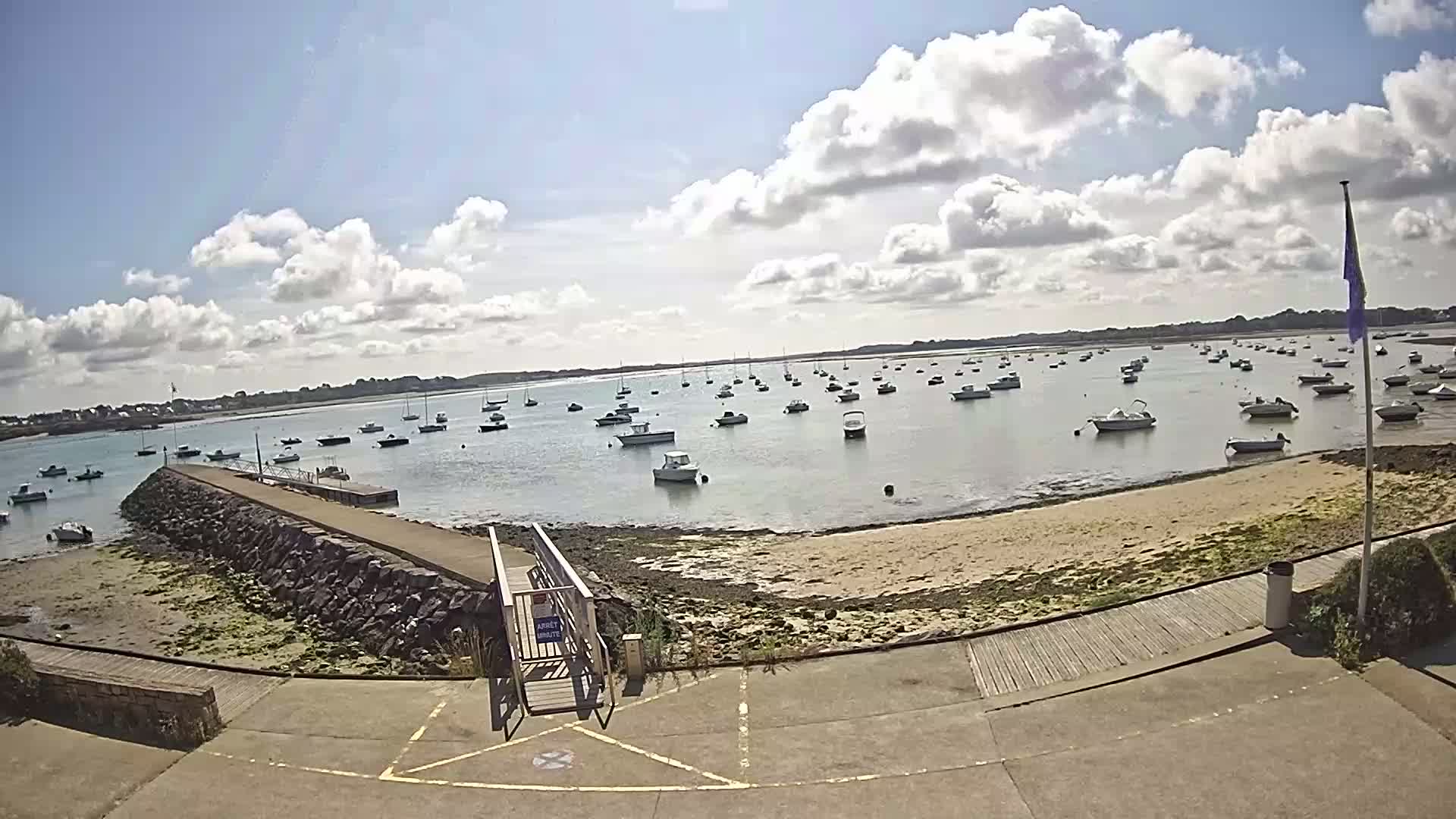 A sunny day with partly cloudy skies shows a calm bay filled with numerous small boats moored near a rocky shoreline with a paved walkway leading to a ramp.