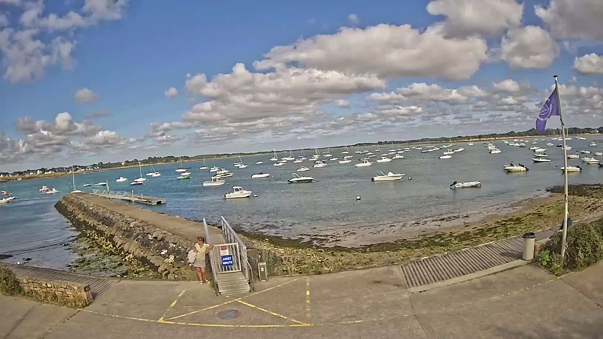 A mostly sunny day with scattered clouds shows numerous boats moored in a calm bay, viewed from a paved area with a stone jetty and a woman standing near a gate.