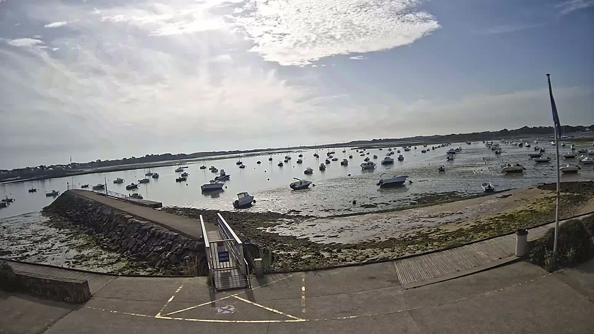 A mostly sunny day reveals numerous small boats resting in a calm harbor at low tide, with a stone jetty and paved walkway leading to the water.