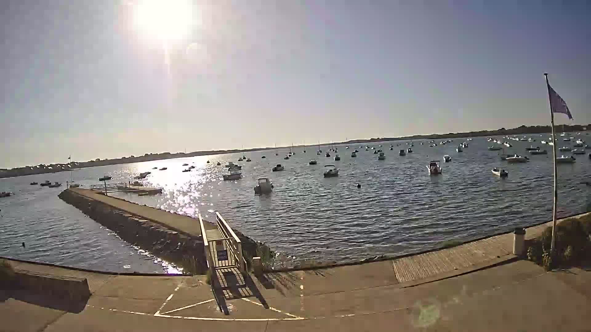 A sunny day shows many small boats moored in a calm bay, viewed from a paved ramp leading to a pier.
