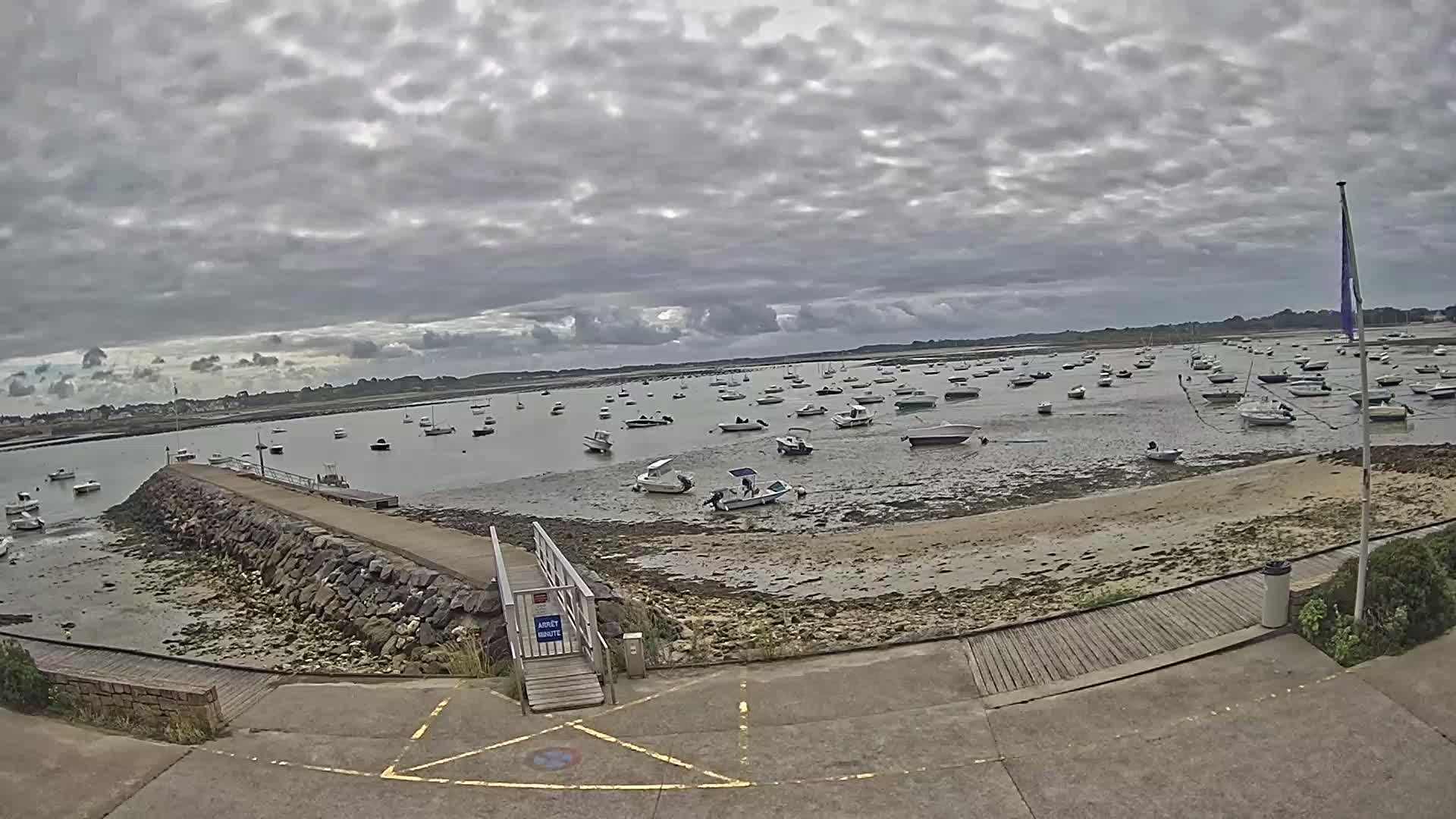 Many small boats are moored in shallow, calm water under a cloudy sky on a sandy shore next to a stone jetty and paved walkway.