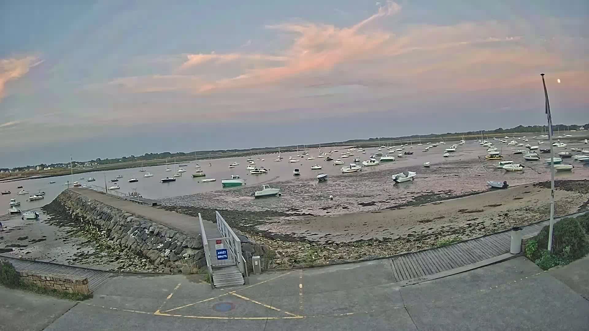 Many boats are moored in a calm harbor at low tide under a partly cloudy, pale pink and blue sky.