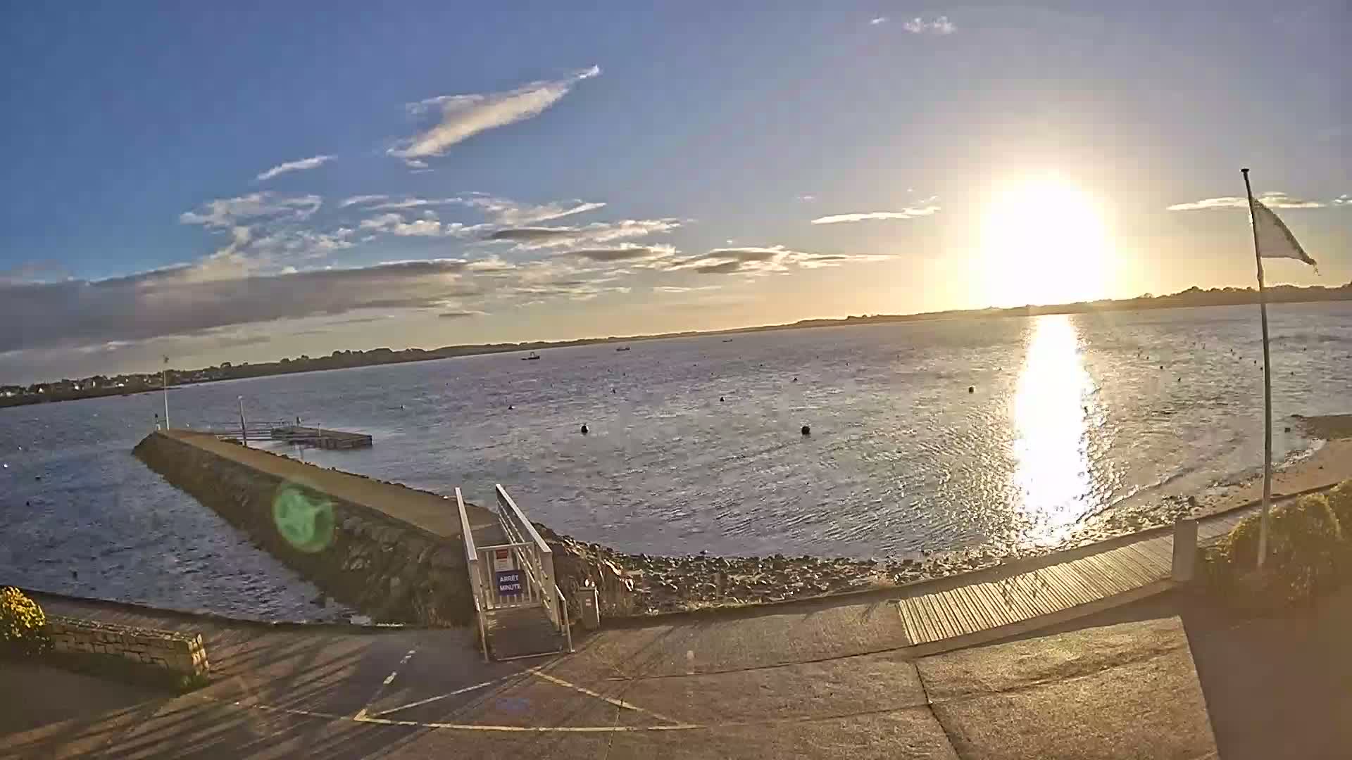 The bright low sun reflects intensely on the rippled waters of a bay featuring a pier, scattered buoys, and distant boats, all under a partly cloudy blue sky.