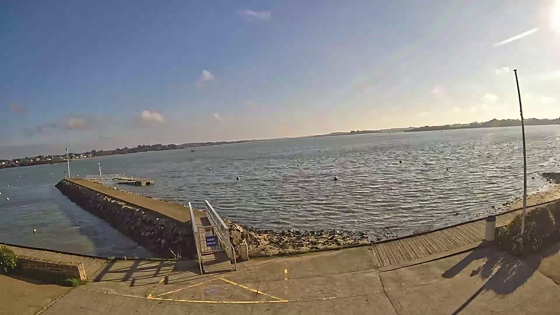 The bright low sun reflects intensely on the rippled waters of a bay featuring a pier, scattered buoys, and distant boats, all under a partly cloudy blue sky.