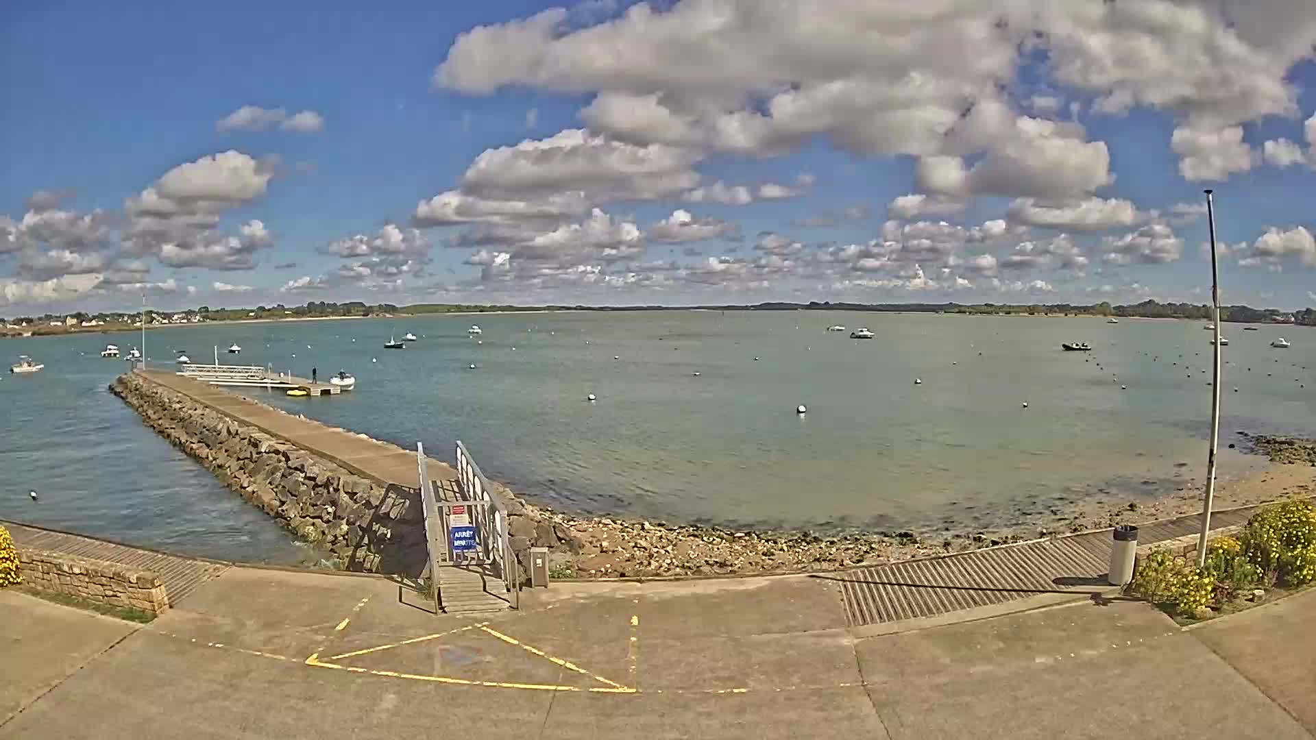 The bright low sun reflects intensely on the rippled waters of a bay featuring a pier, scattered buoys, and distant boats, all under a partly cloudy blue sky.
