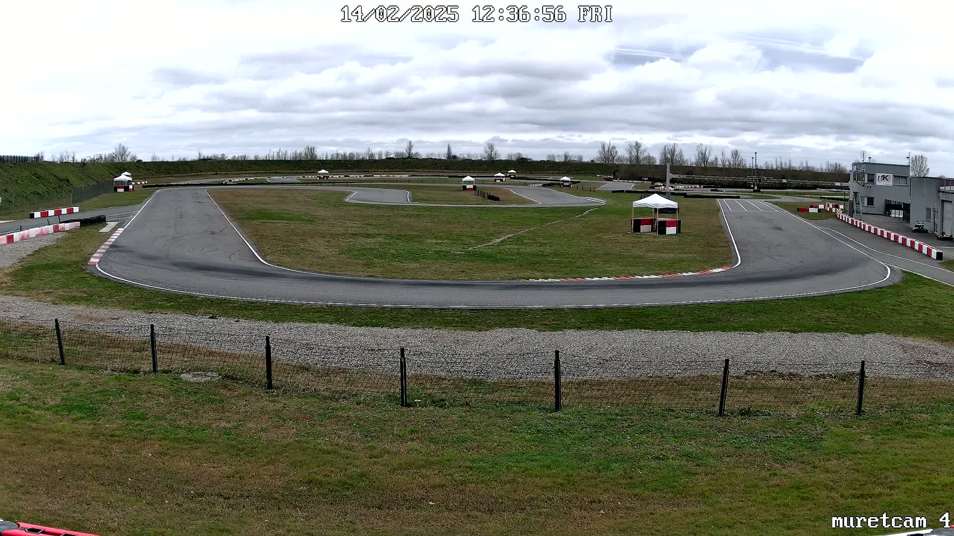 An outdoor go-kart track with overcast skies is seen from a slightly elevated viewpoint, showing the asphalt track winding through grassy areas with small white tents and red-and-white barriers.
