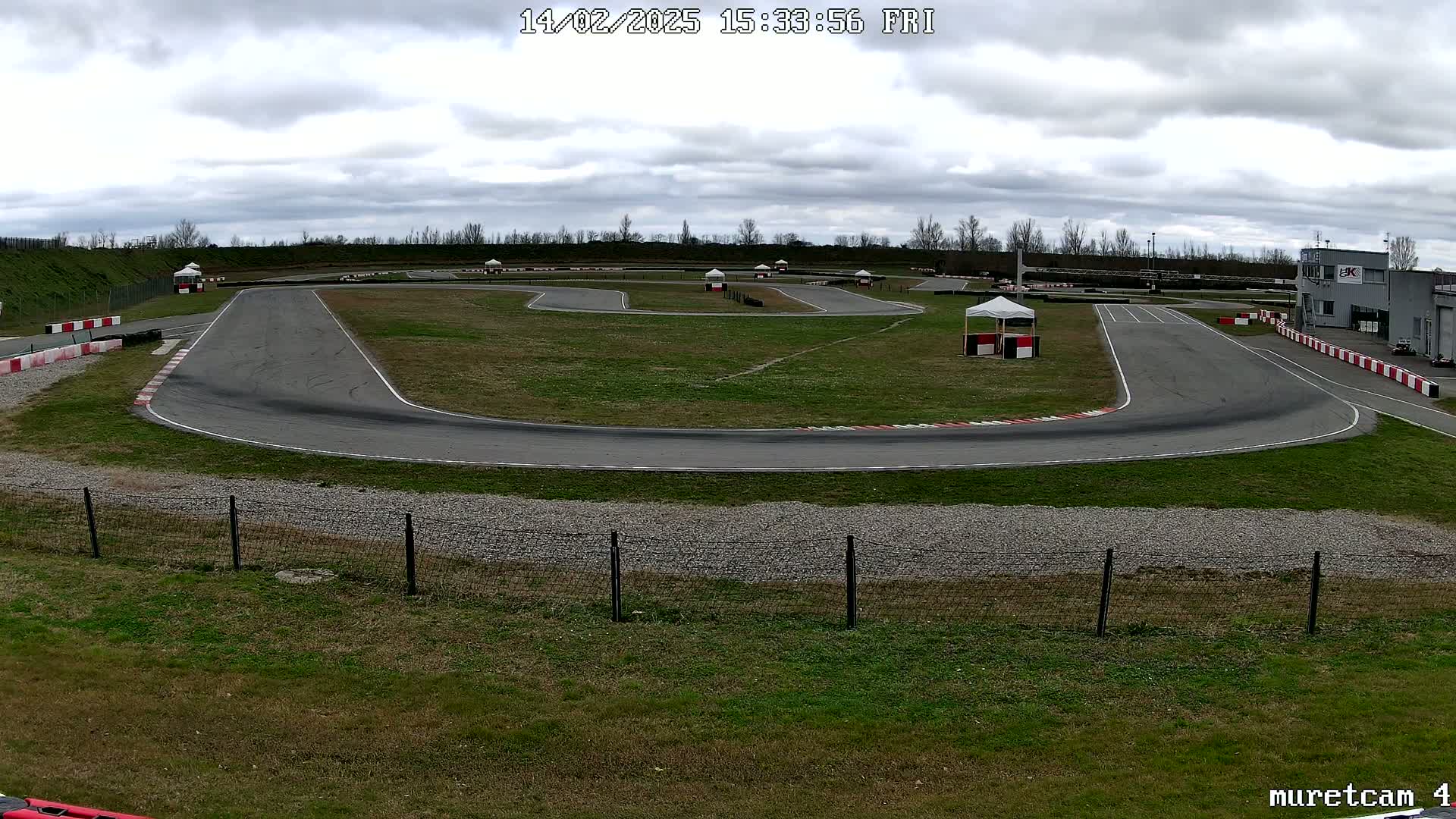 An outdoor go-kart track with overcast skies curves through a grassy field, bordered by fencing.