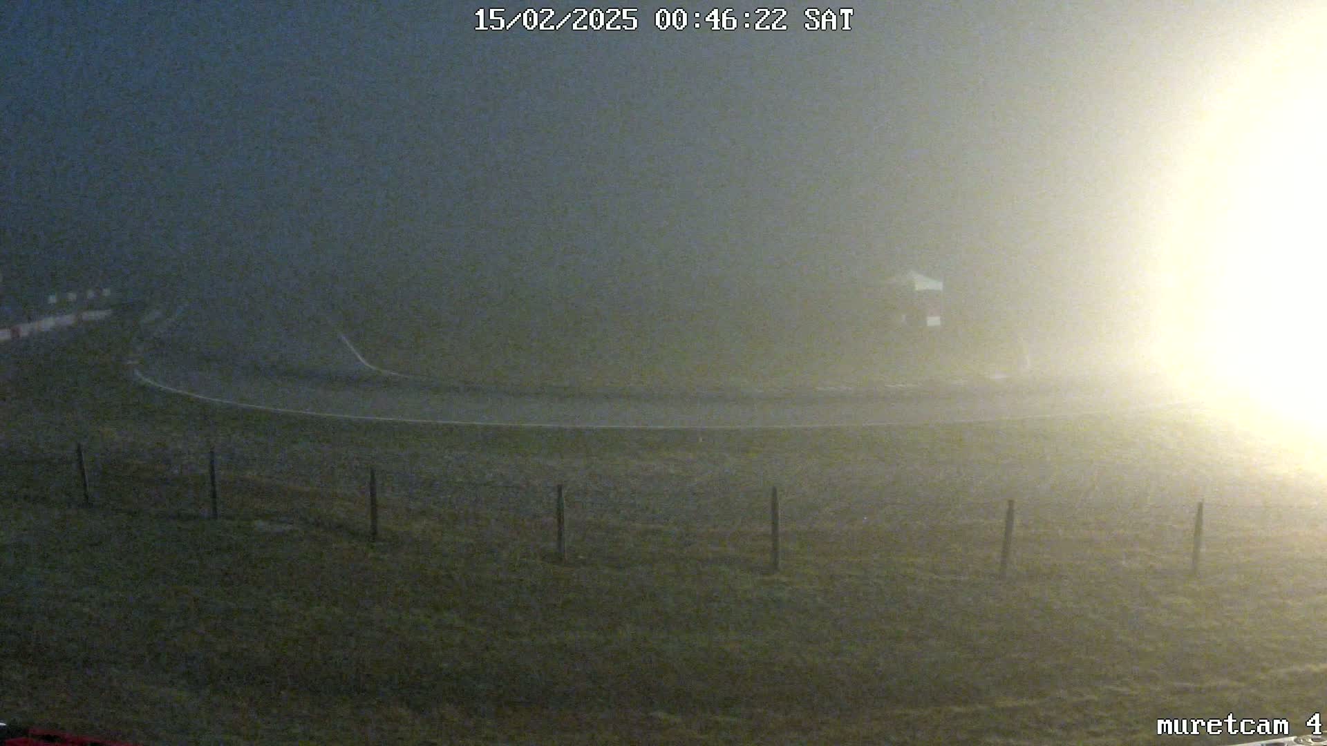 A foggy, dimly lit outdoor scene shows a curved roadway and a fence in the foreground, with a building barely visible in the mid-ground.