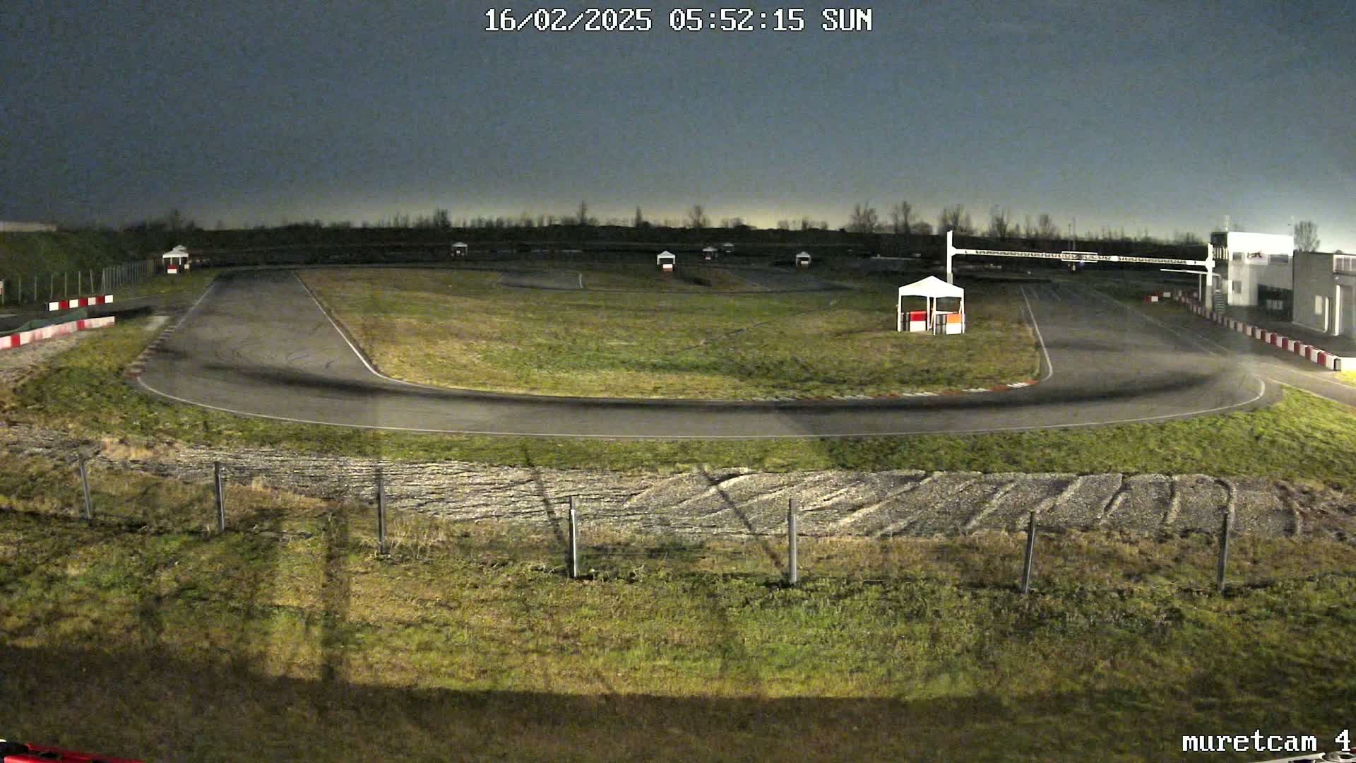 An outdoor go-kart track at night under a dark sky, showing a paved track winding through grassy areas, with small structures and fencing visible.